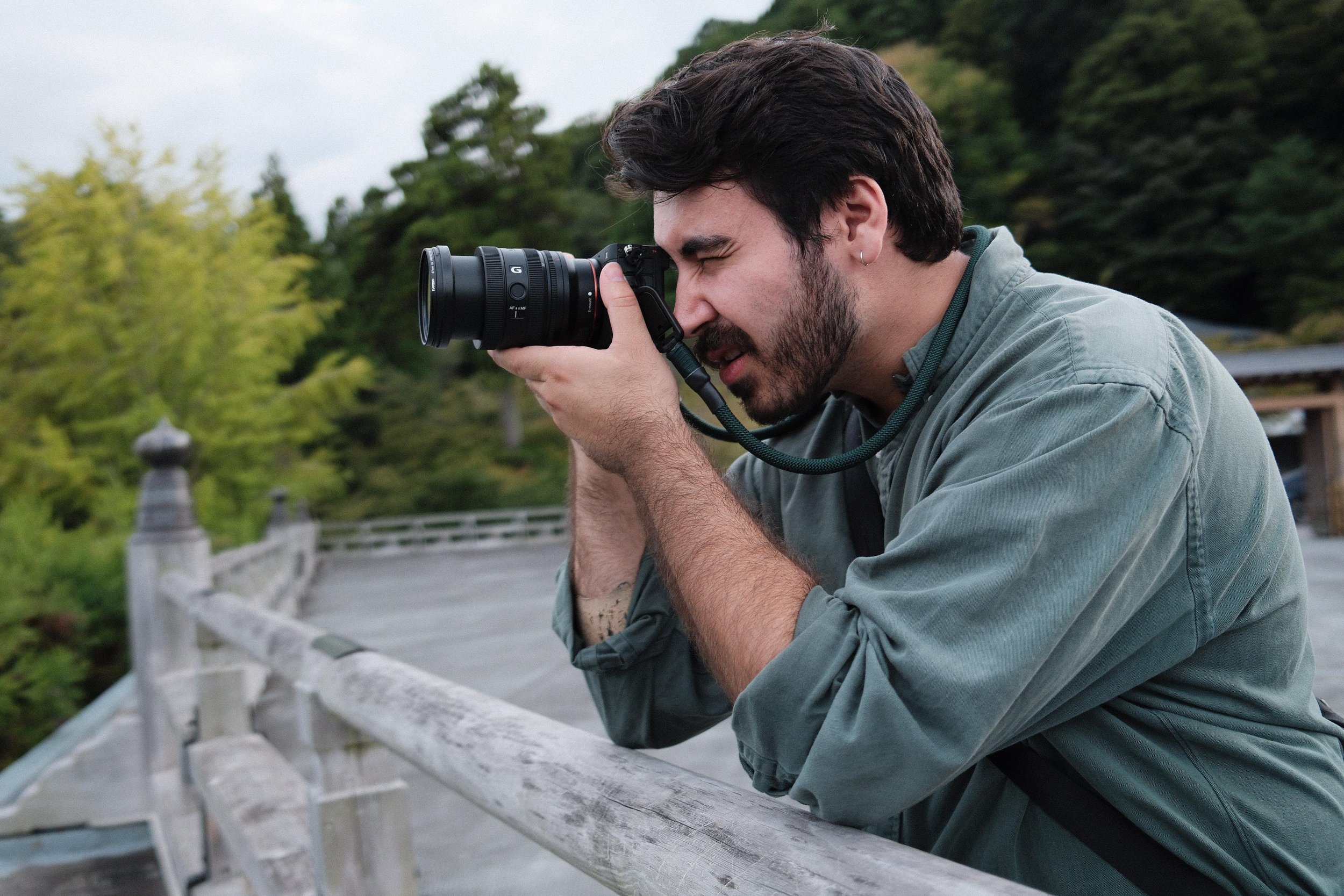 A man with dark hair and a beard taking a photograph with a DSLR camera on a wooden bridge surrounded by green trees.