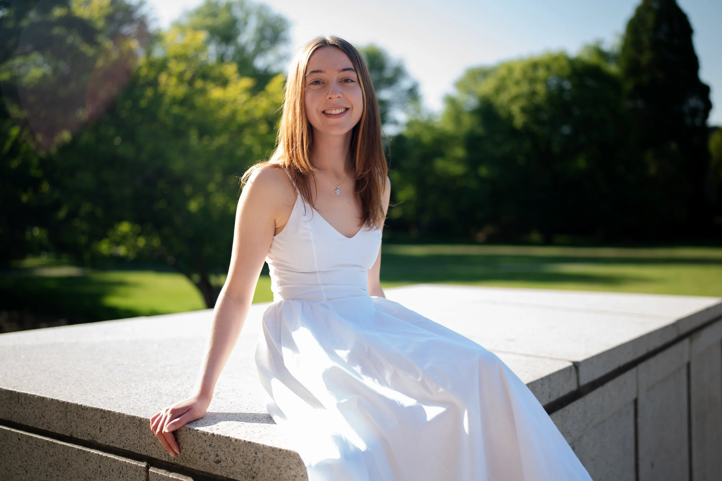 A young woman in a white dress sitting on a concrete ledge outdoors with trees and a grassy park in the background, smiling.