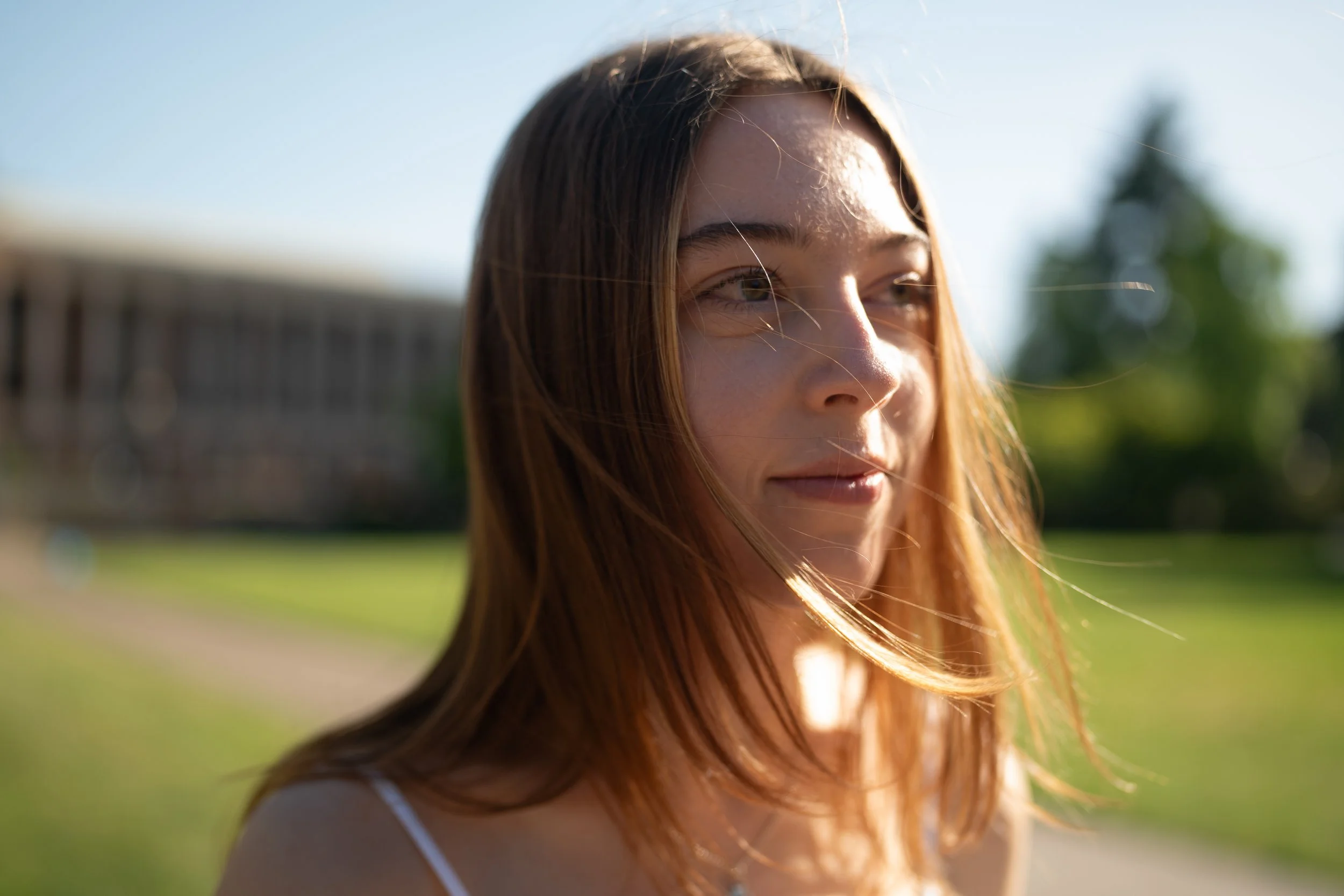 A young woman with long brown hair smiling outdoors on a sunny day, with a blurred background of green grass and trees.