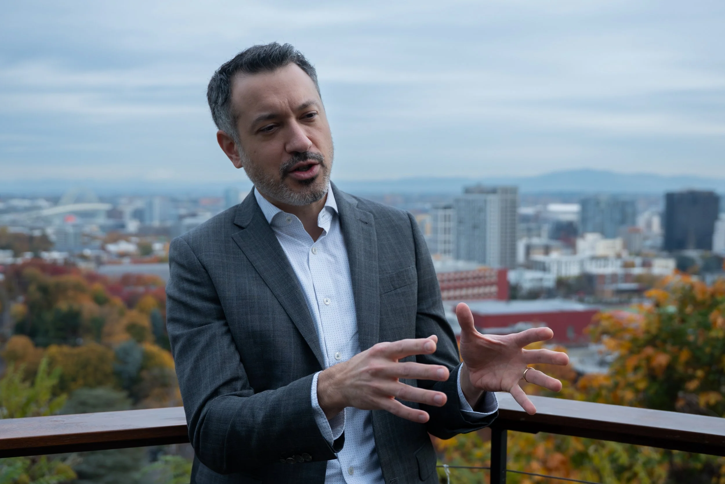 A middle-aged man in a gray suit and white shirt speaking and gesturing with his hands on a balcony overlooking a city skyline with fall foliage.