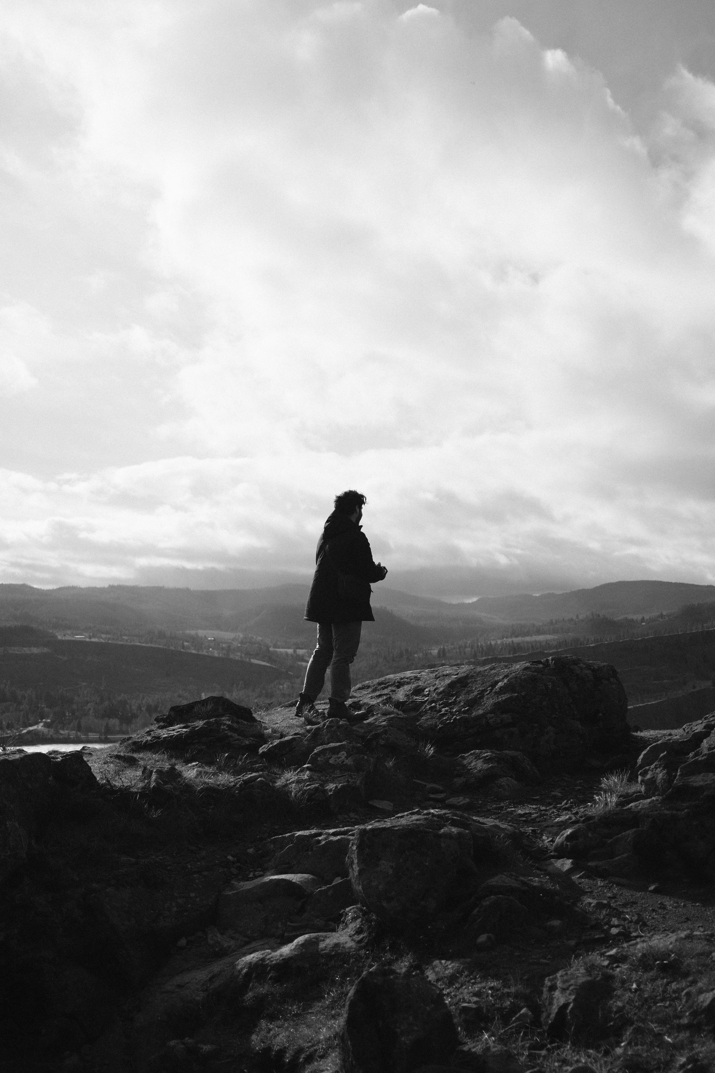 A person standing on rocks overlooking a landscape of hills, with cloudy sky above, in black and white.