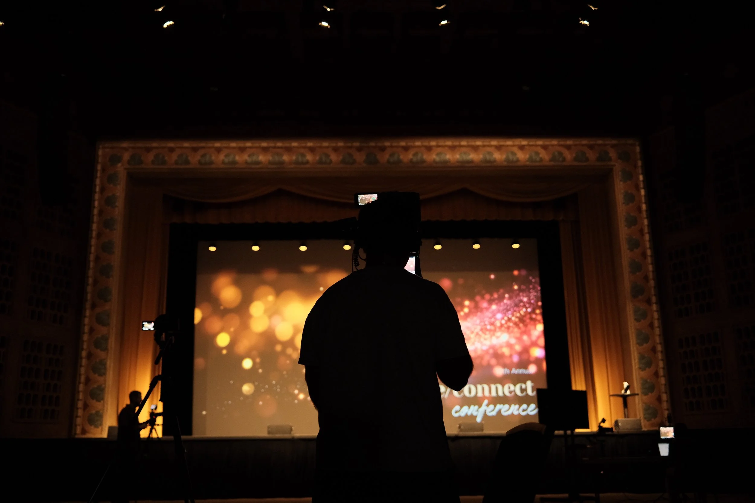 A person taking a photo of a stage with a large screen displaying '2023 Connect Conference' and colorful bokeh background, with other equipment and crew members in the dark.