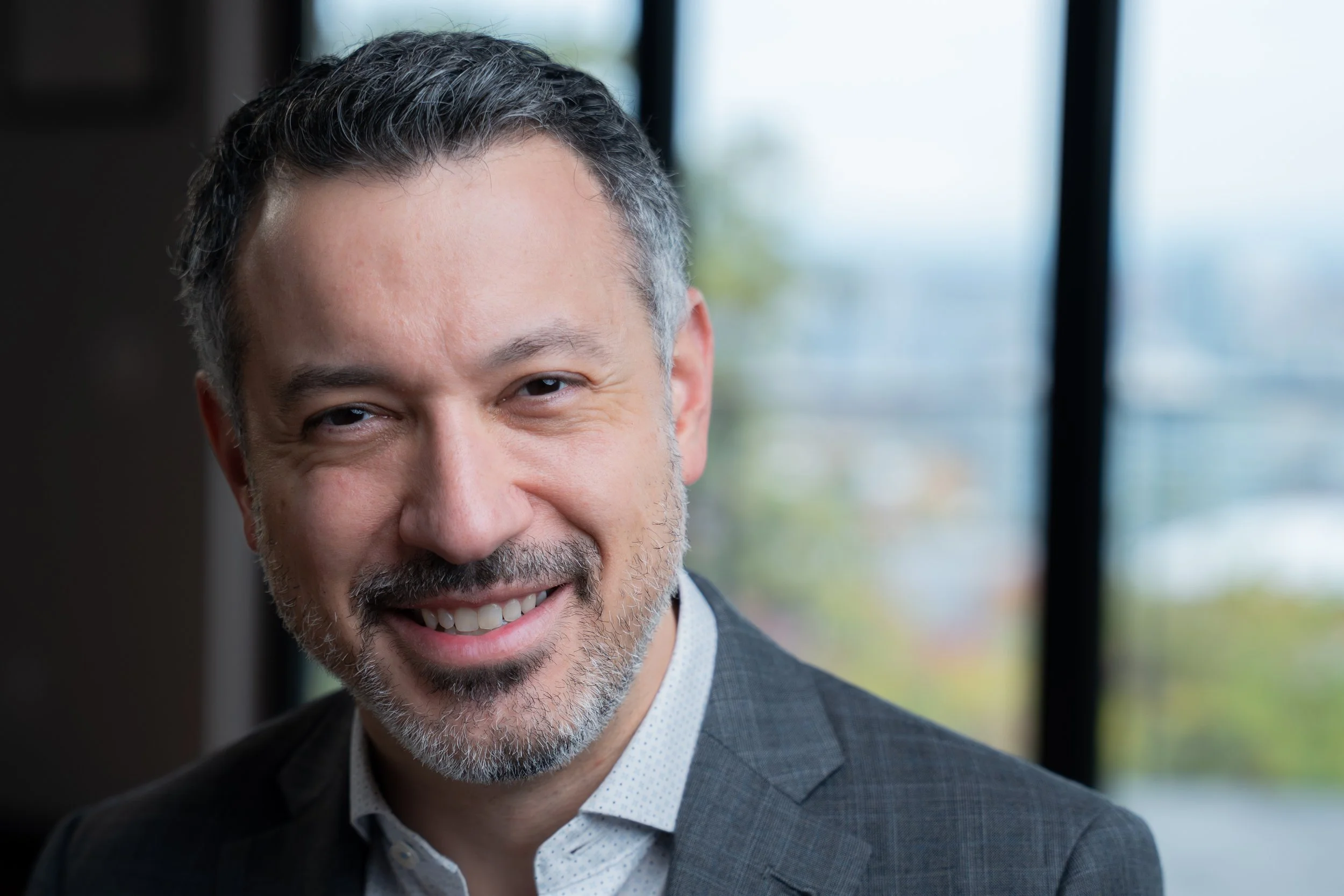 Close-up of a smiling middle-aged man with gray hair and a beard, wearing a gray suit jacket and white shirt, standing indoors with windows in the background.