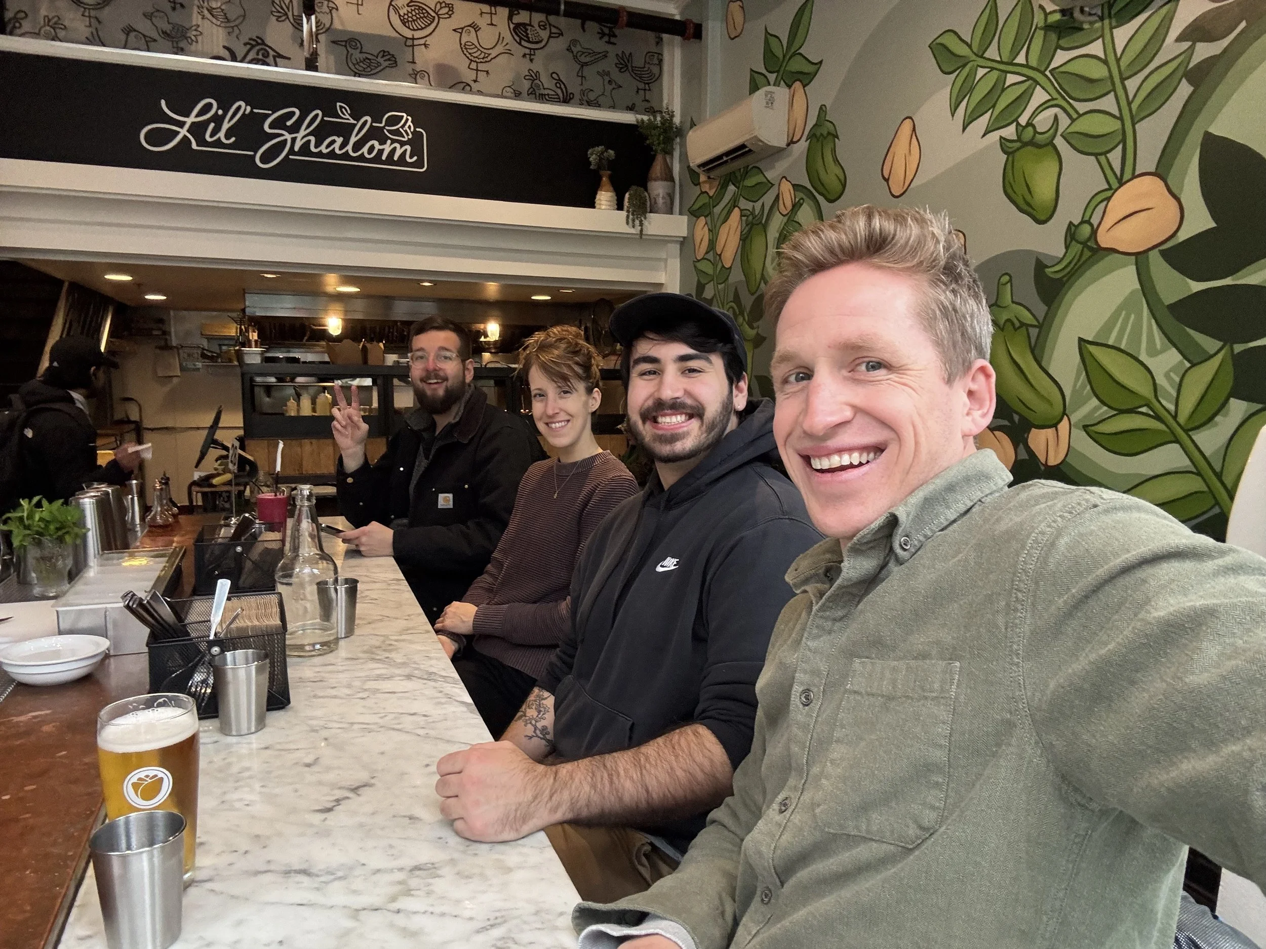 Group of five friends smiling and taking a selfie at a restaurant counter with a mural of vegetables on the wall behind them.