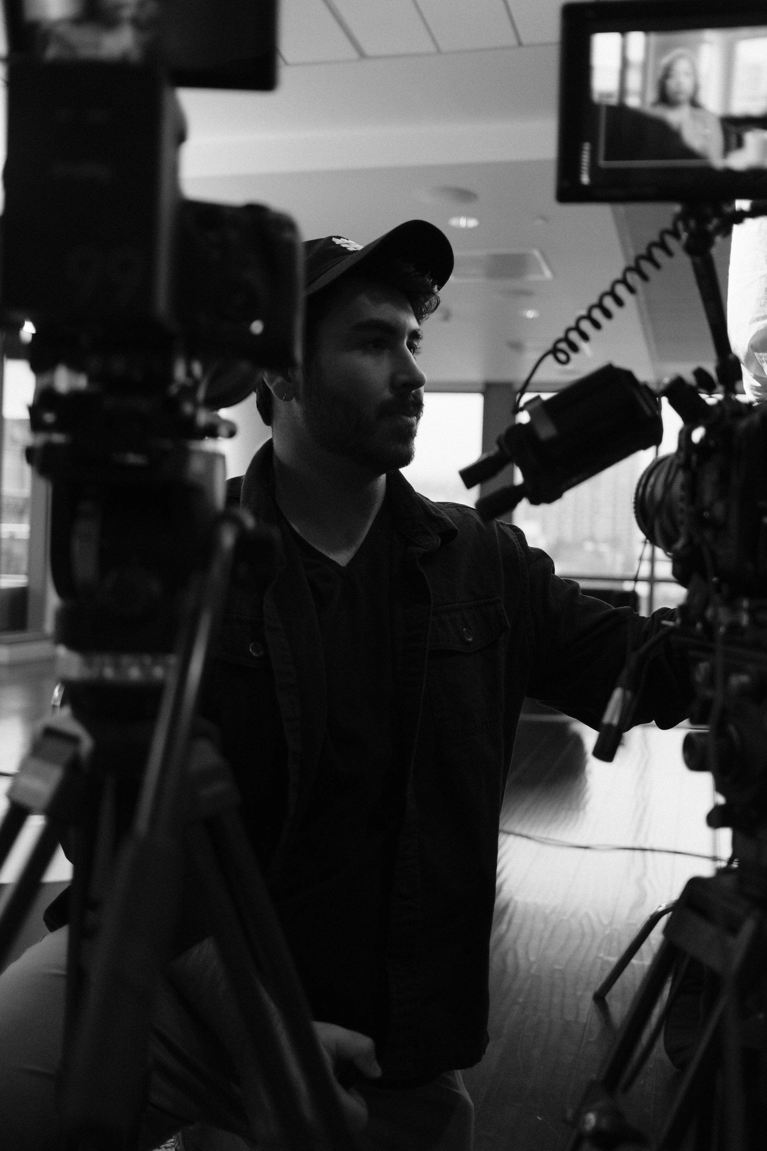 A man in a baseball cap operating a professional video camera indoors with large windows in the background.