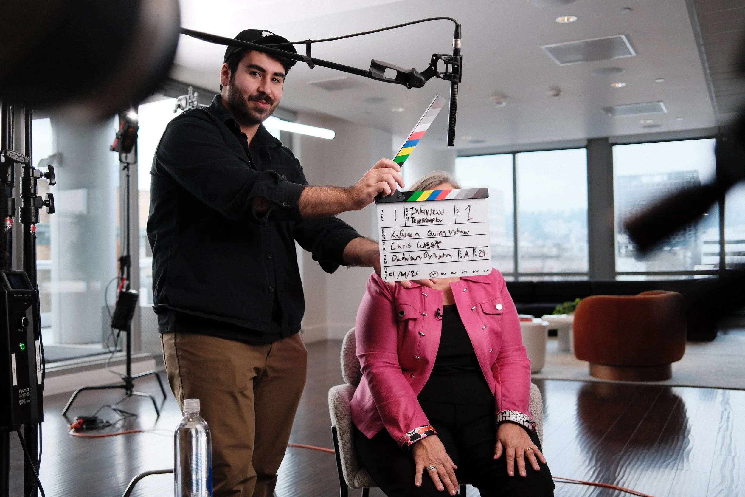Filming scene with a man holding a clapperboard and a woman seated, in a modern indoor setting with large windows