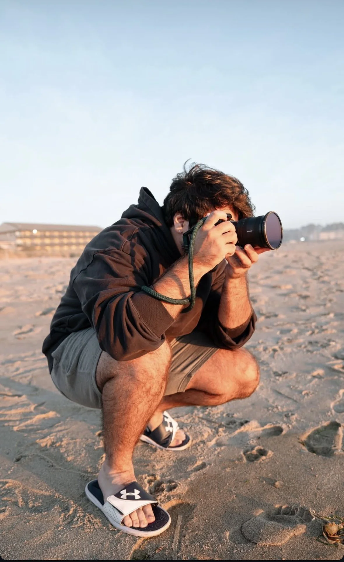 A man crouching on a sandy beach, taking a photo with a camera, wearing a dark hoodie, shorts, and flip-flops during sunset or sunrise.