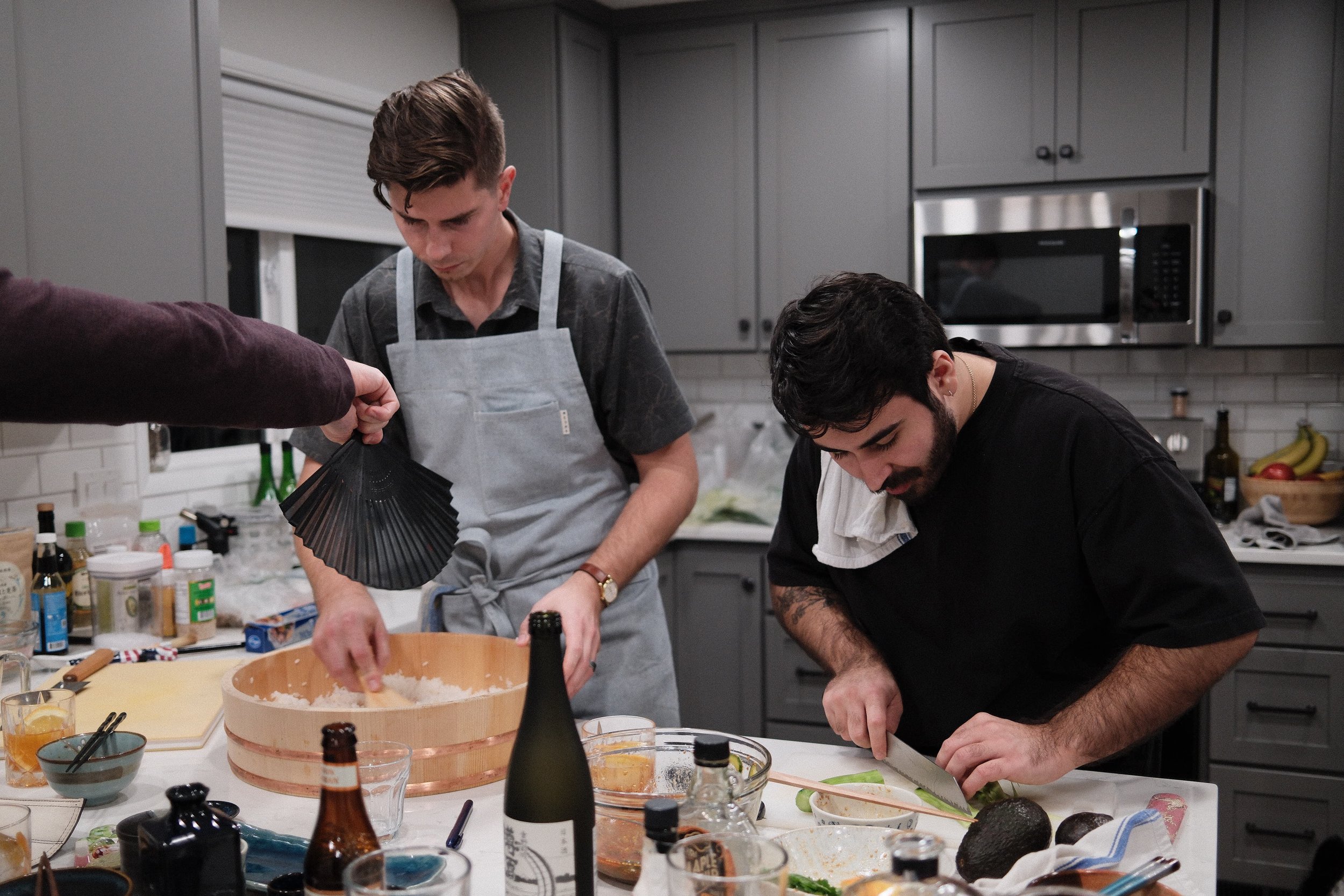 Two men preparing food in a modern gray kitchen, one is slicing an avocado and the other is mixing ingredients in a bowl, with various bottles and food items on the countertop.