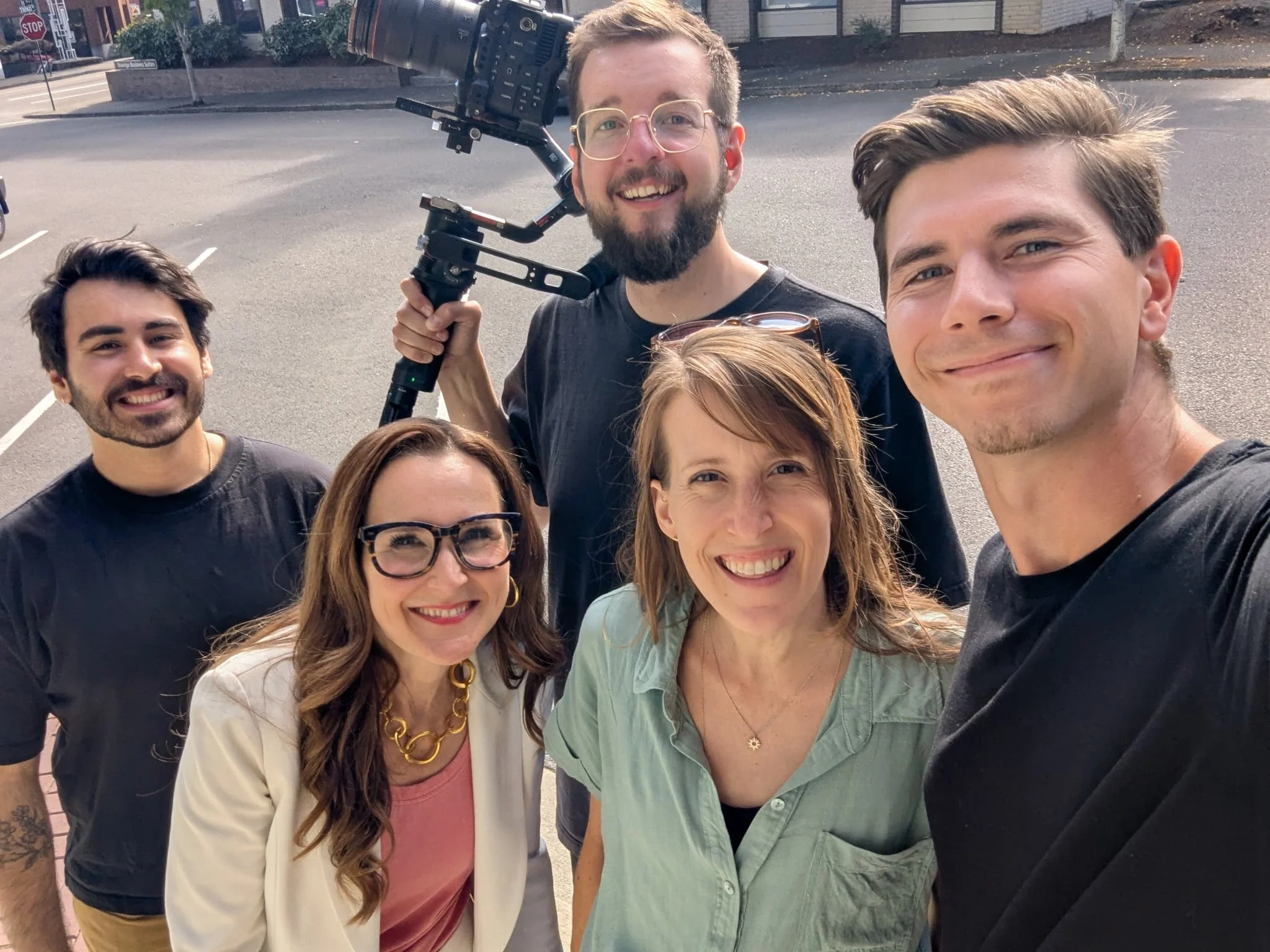 Group of six people smiling for a selfie outdoors, with one person holding a camera on a stabilizer.