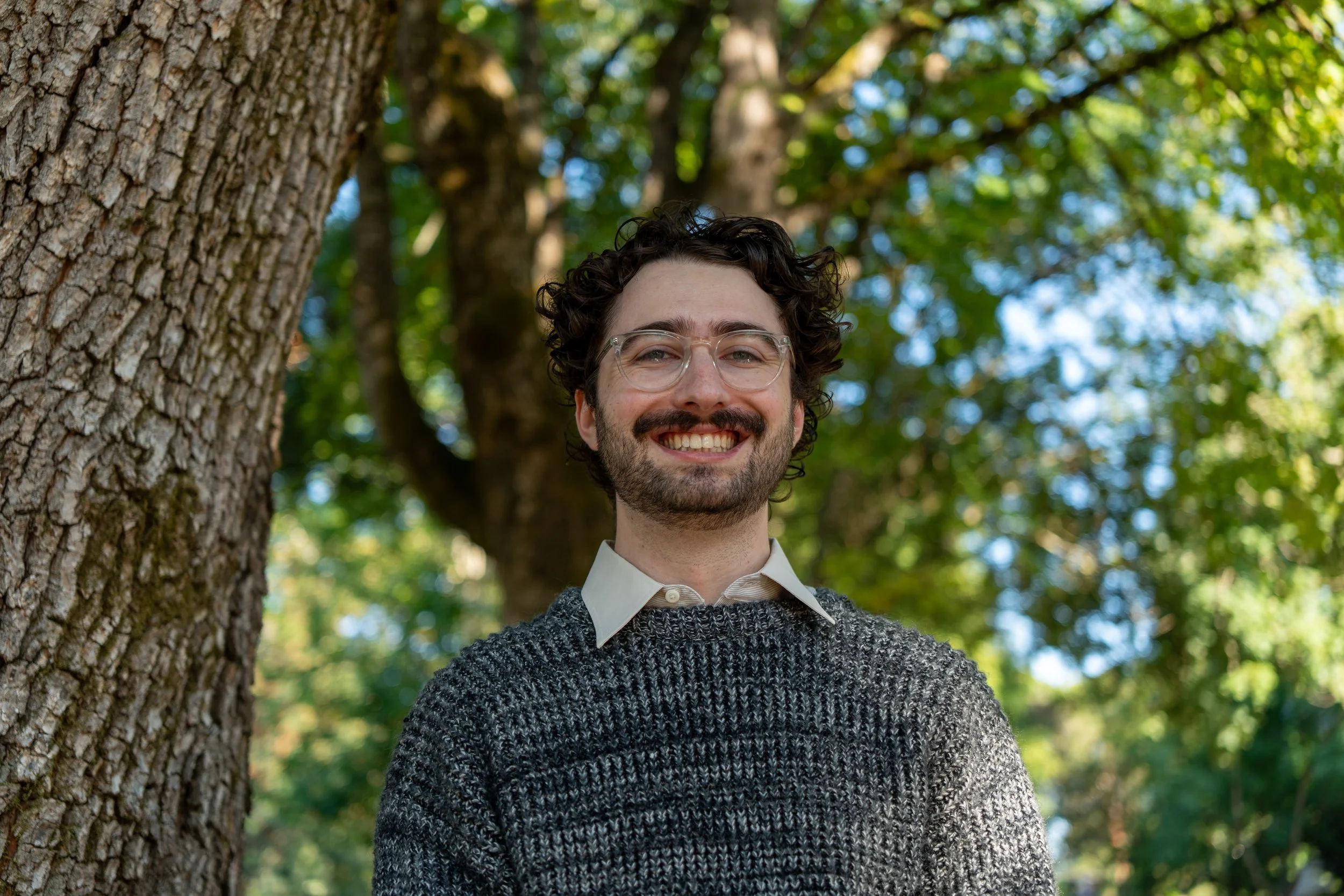 A smiling man with curly dark hair, glasses, and a beard, standing outdoors in front of a large tree with green leaves.