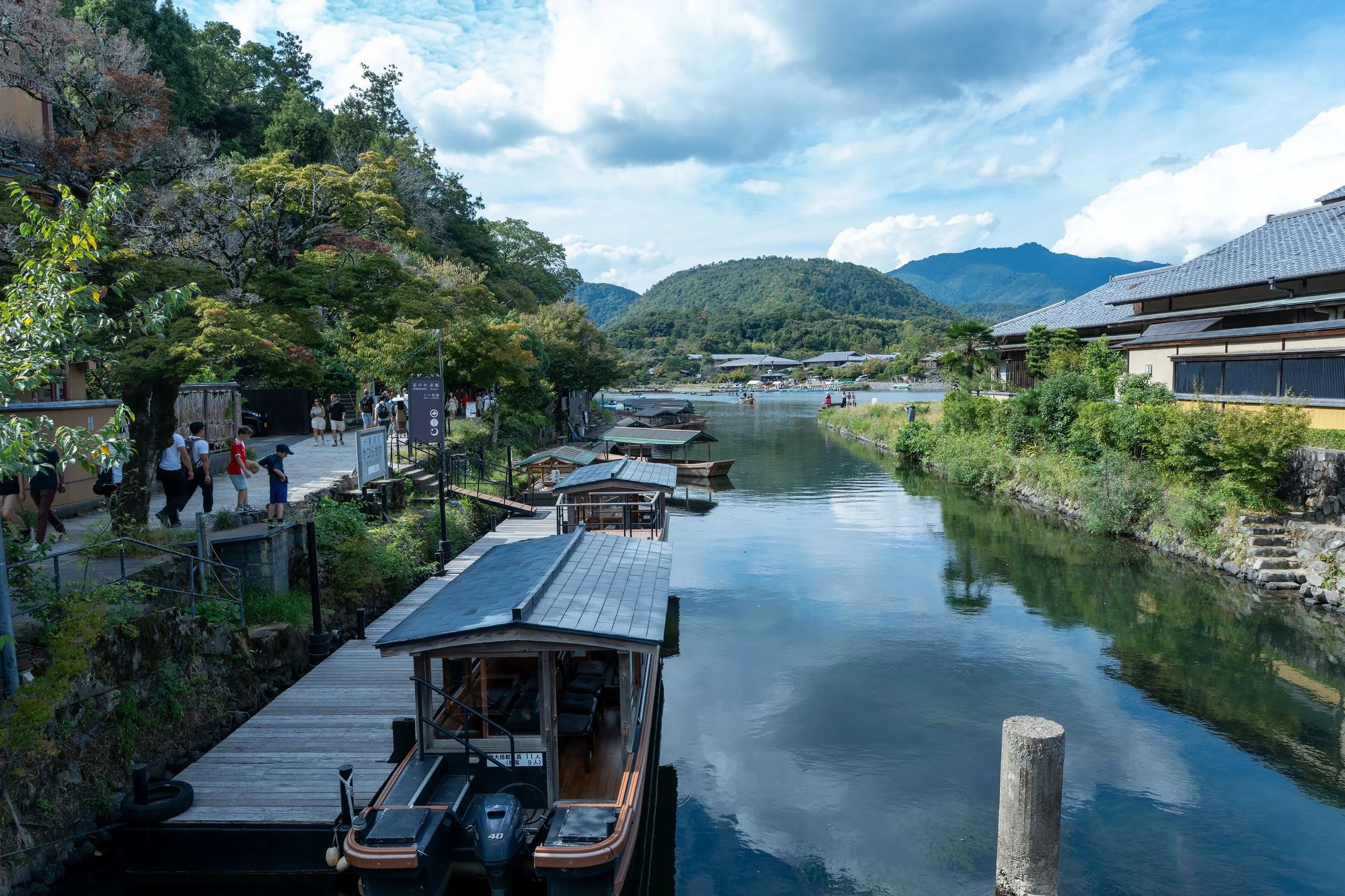 A scenic view of a river with boats docked along a wooden pier, surrounded by lush green trees, traditional buildings, and distant mountains under a partly cloudy sky.