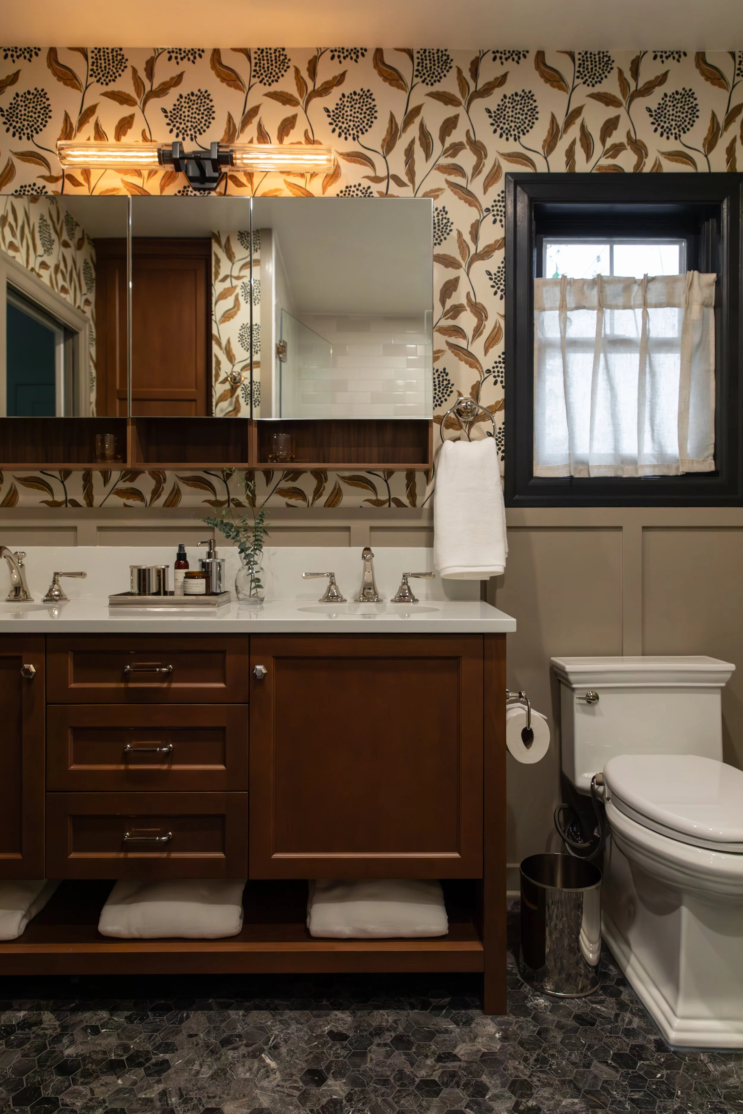 A bathroom with a wooden vanity, a mirror, a small window with a curtain, and a toilet. The wall has a floral wallpaper and a mirror cabinet with open shelves. The sink has a faucet, some toiletries, and a small plant. There are shelves beneath the v