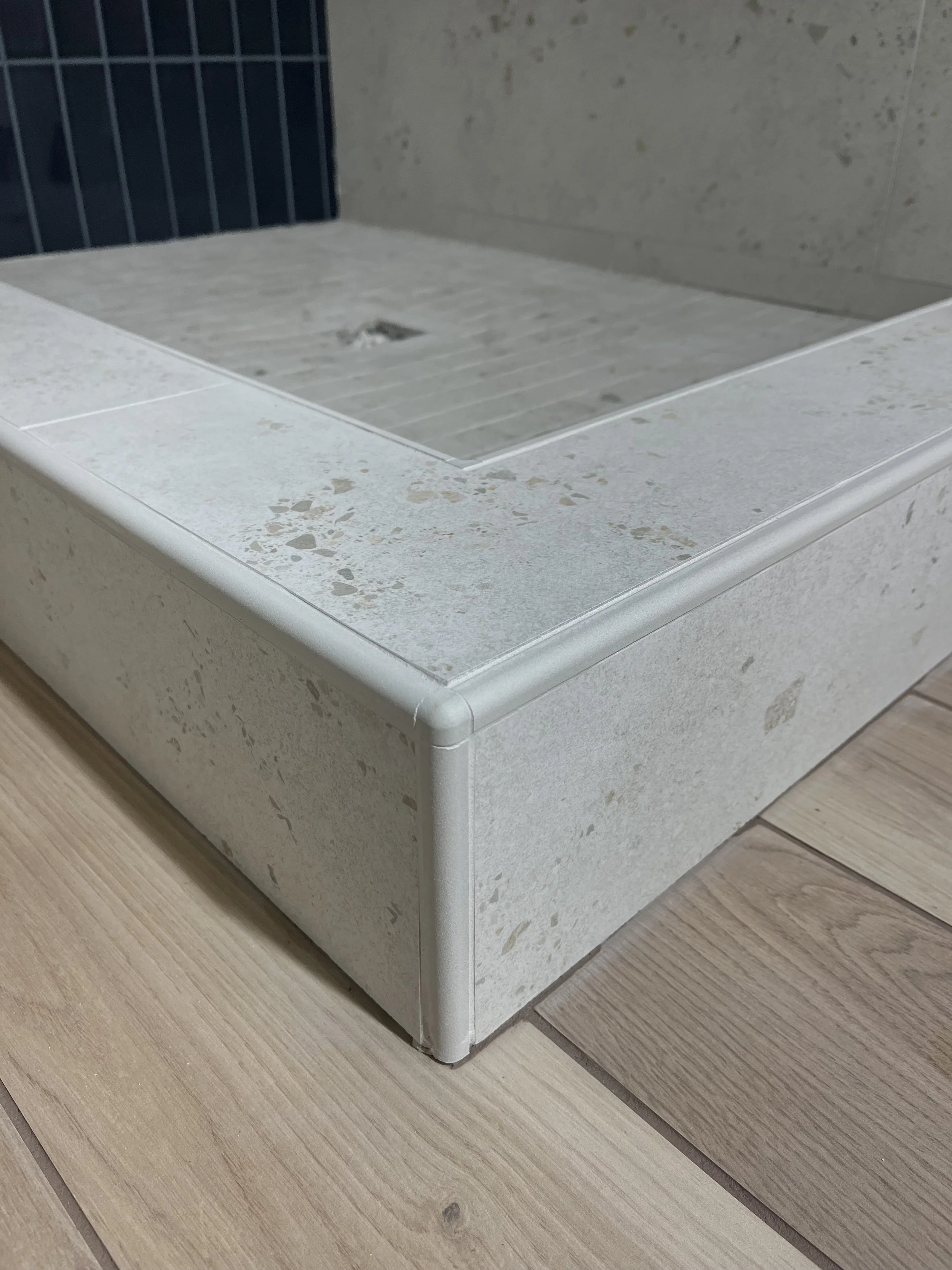 Close-up of a modern, rectangular, beige stone shower curb with a gray tiled interior and a rounded white edge, situated on a wooden floor.
