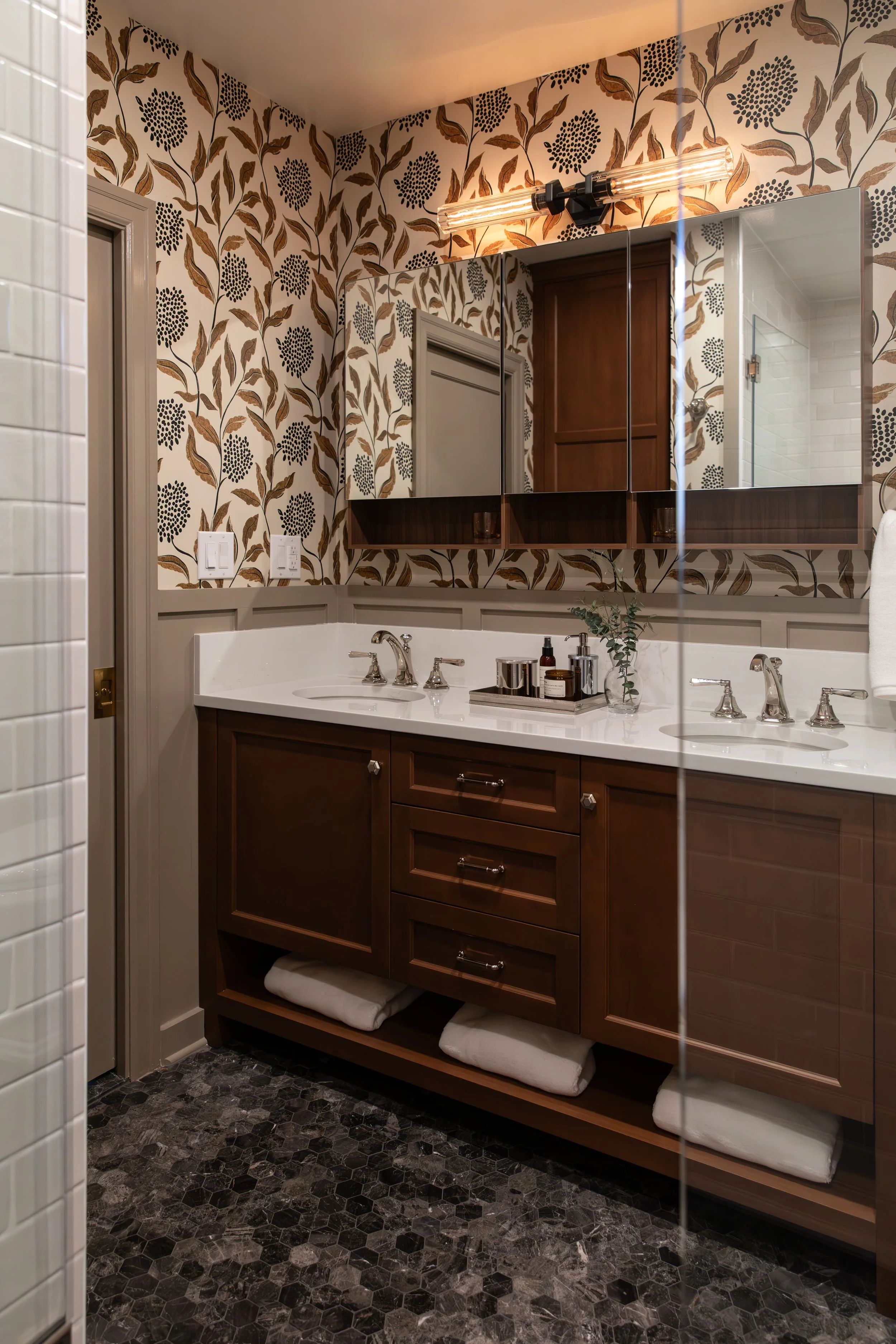 A bathroom vanity with a white countertop, dual sinks with silver faucets, brown wooden cabinets, and a large mirror. The wall has floral-patterned wallpaper, and there are neatly folded white towels underneath the vanity. Small toiletries and a plan