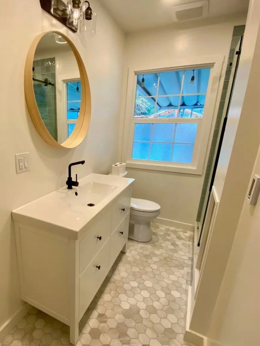 A small bathroom with a white vanity, black faucet, toilet, hexagonal tile floor, a round mirror, vanity light fixtures, a window with a view of a blue canopy outside, and a partially visible shower with green tile.