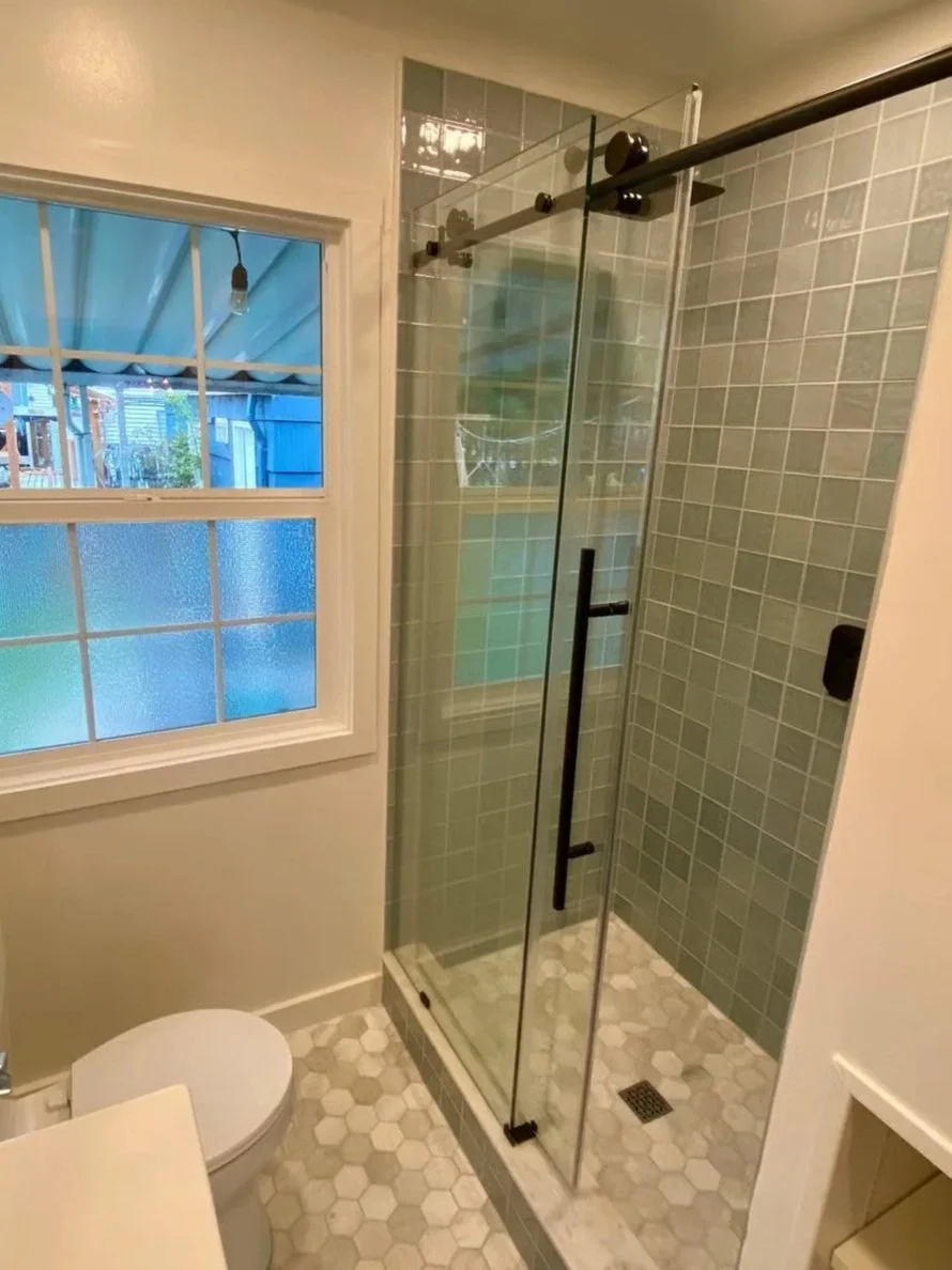 A small bathroom with stone hexagon tile flooring, a frosted window, a toilet, and a glass shower enclosure with green tiles inside.