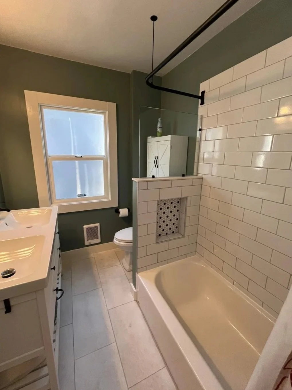 Small bathroom with a green painted wall, white tile accents, a window, a white vanity with dual sinks, a toilet, and a shower with white subway tiles and a decorative niche.
