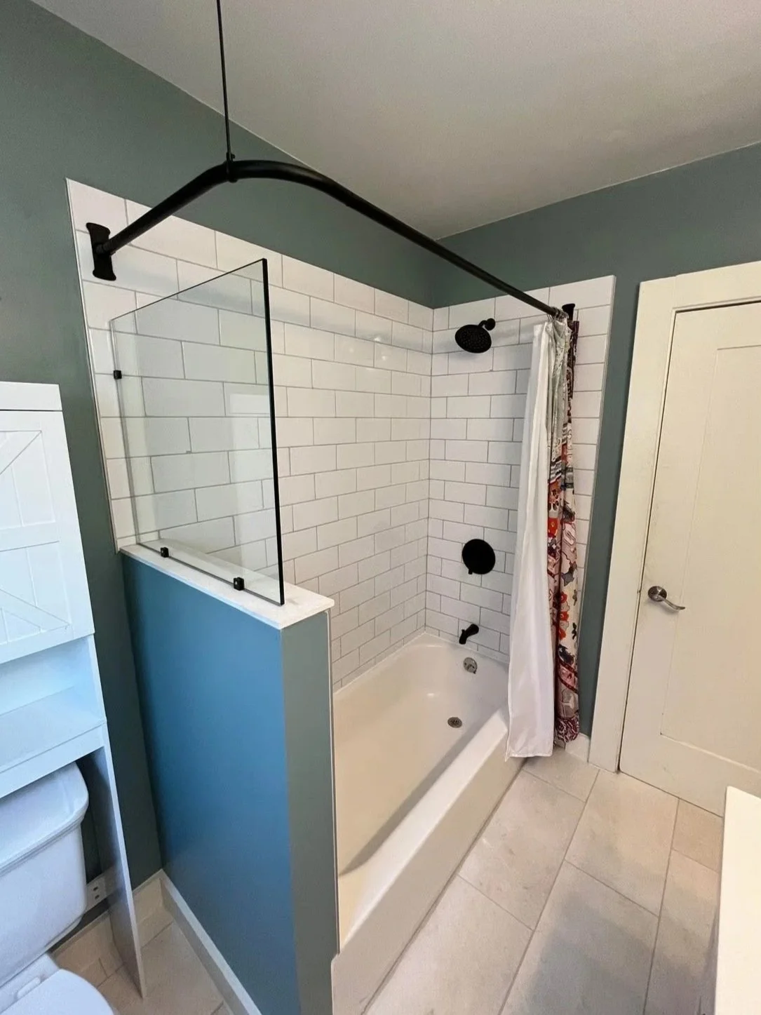Bathroom shower with a black showerhead and fixtures, white subway tile walls, a partial glass shower divider, a white bathtub, a colorful shower curtain, a beige tile floor, and a closed white door.