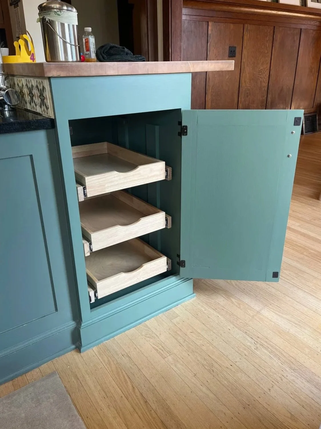 Open teal-colored cabinet with three empty wooden pull-out shelves, set in a kitchen with wood flooring and a countertop with various items on top.