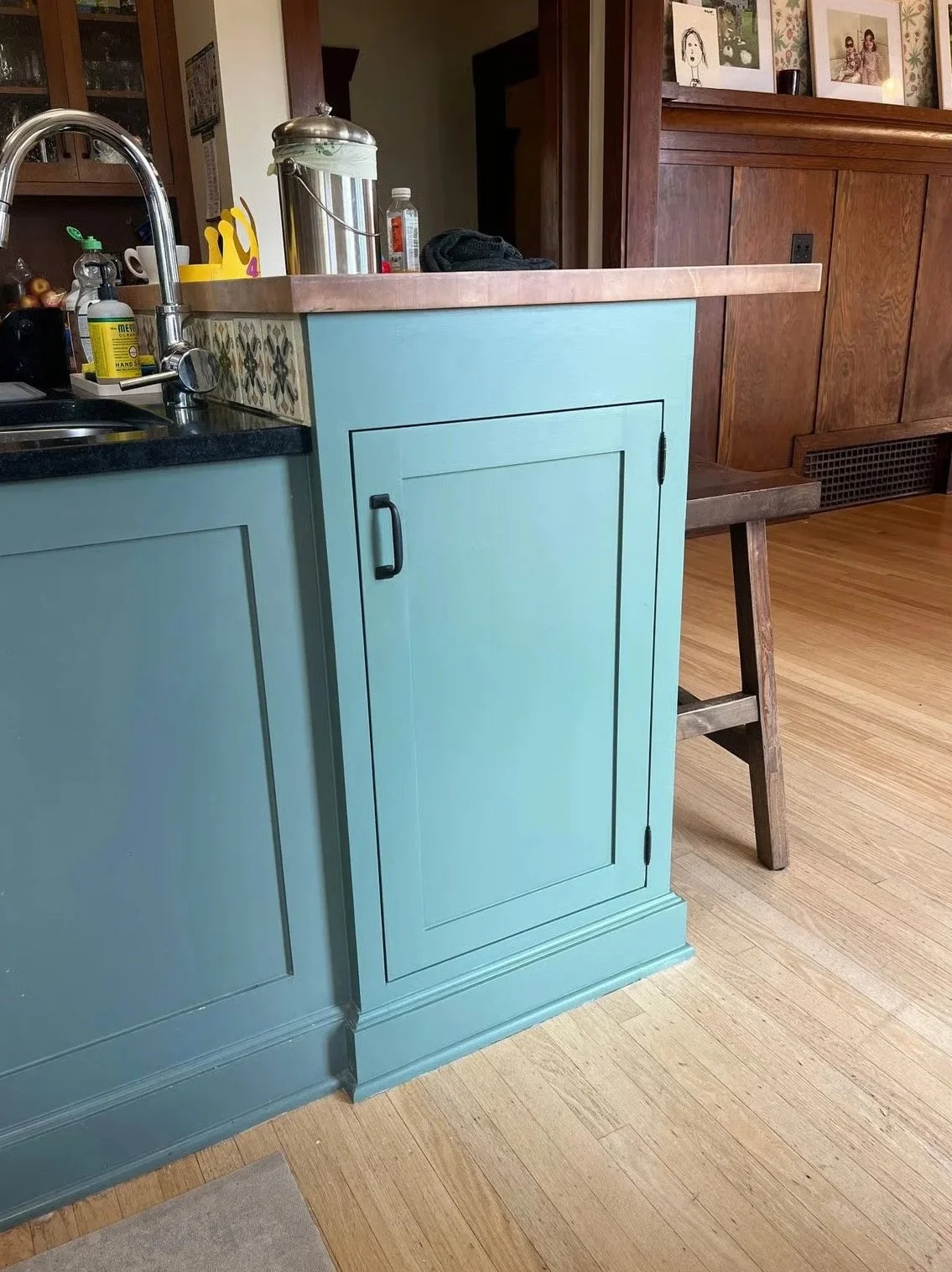 A kitchen with blue cabinets, a black countertop, a sink, and a cutting board. There are items on the counter, including a yellow utensil, a bottle of hand sanitizer, and a silver container. A wooden chair is beside the cabinets, and the floor is har