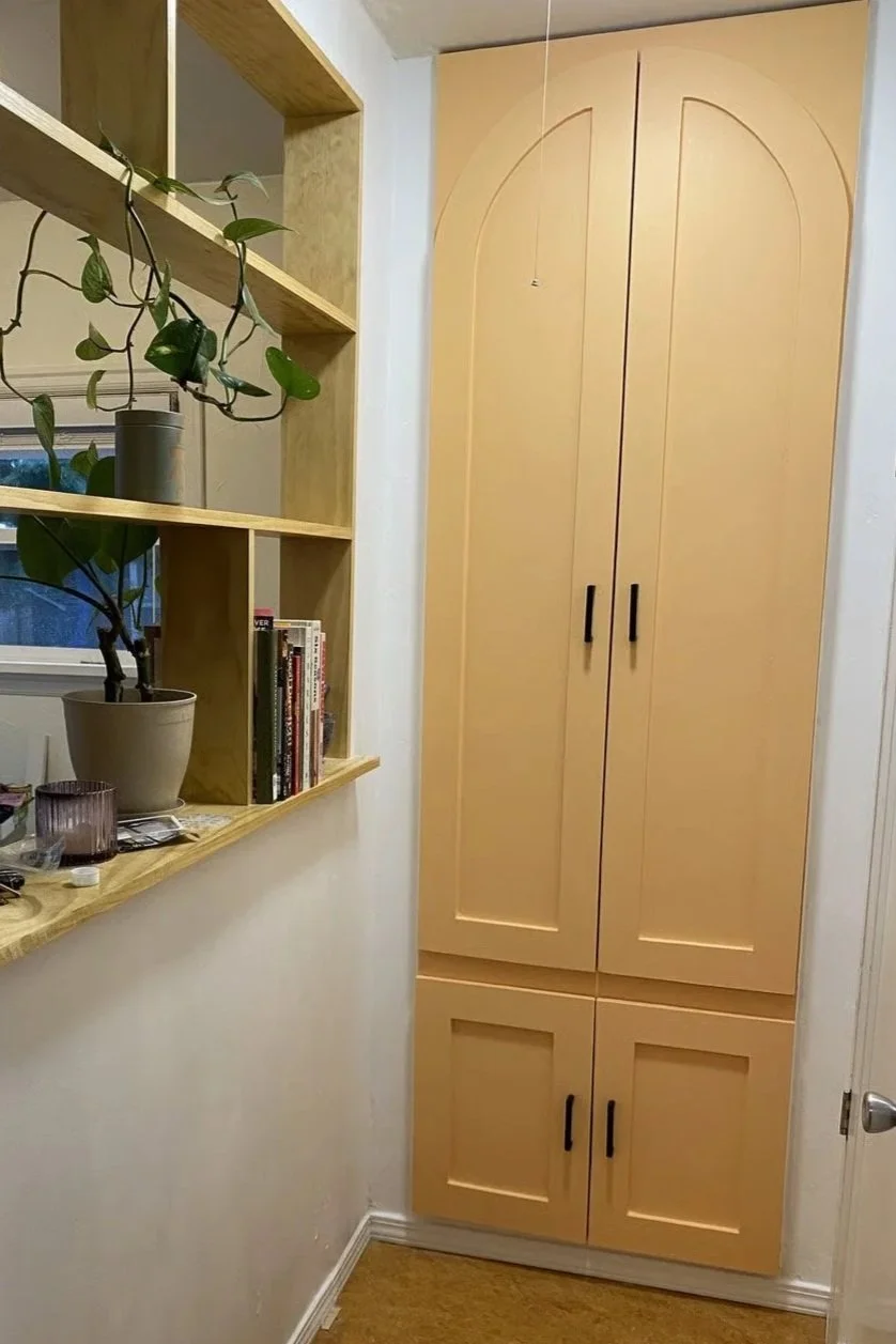 A built-in, double-door wooden cabinet in a small room or hallway, next to a white wall. To the left, there is a light-colored wooden shelf with a potted plant, some books, and other miscellaneous items. A window with a partially open white blind is 