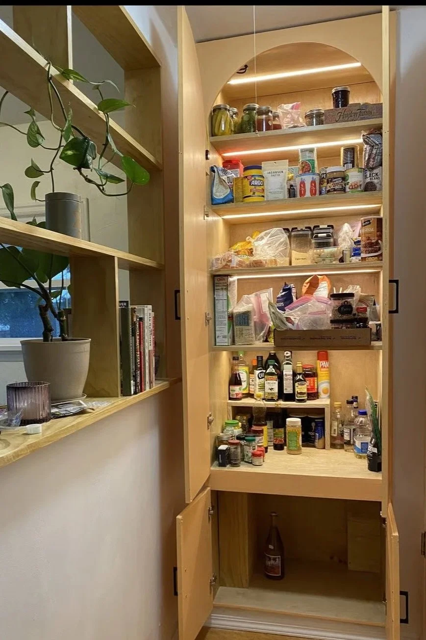 A tall wooden kitchen pantry with open doors containing various jars, bottles, and food items on shelves, and a bottle on the bottom shelf.