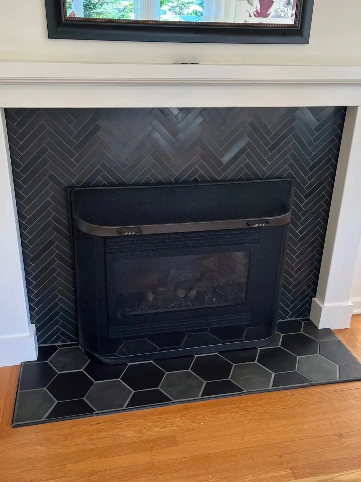 Black fireplace insert with a decorative vent, installed inside a fireplace cavity with black tile herringbone pattern, on a maple wood floor, with a white surround and framed artwork above.