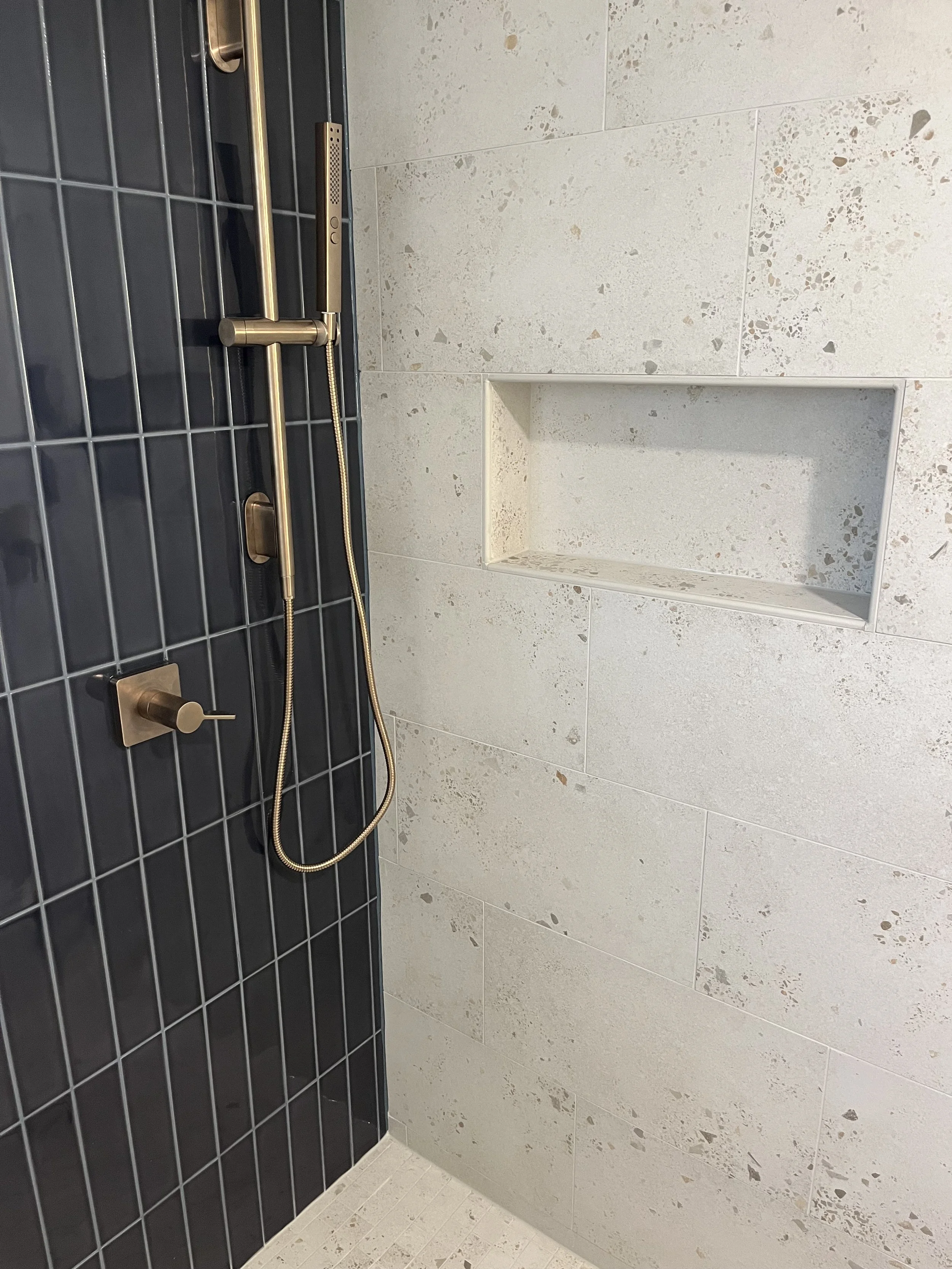 Shower with dark blue tiles, brass fixtures, a built-in niche for toiletries, and a handheld showerhead.