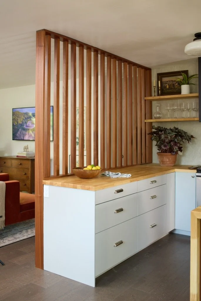 A kitchen area with white cabinets, wooden countertop, and a decorative wooden slat partition. There are potted plants and shelves with glassware and framed artwork on the wall.