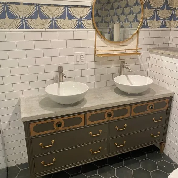 Bathroom with a double vanity, two vessel sinks, and a round mirror. The vanity is dark with gold handles and a gray countertop. The wall has white subway tiles and decorative wallpaper above.