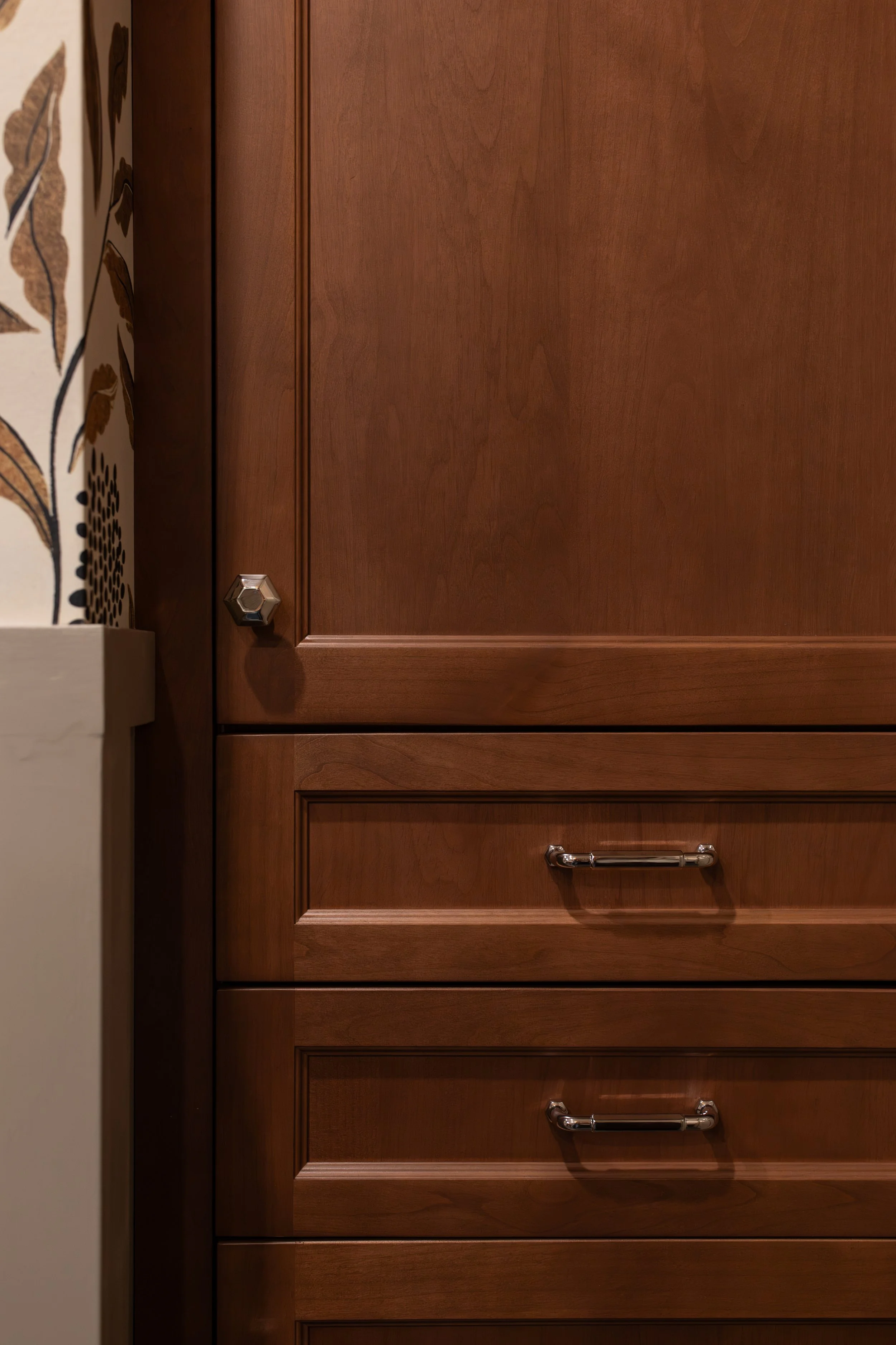 Close-up of a wooden kitchen cabinet with metal handles, next to a wall with patterned wallpaper featuring leaves and plants.