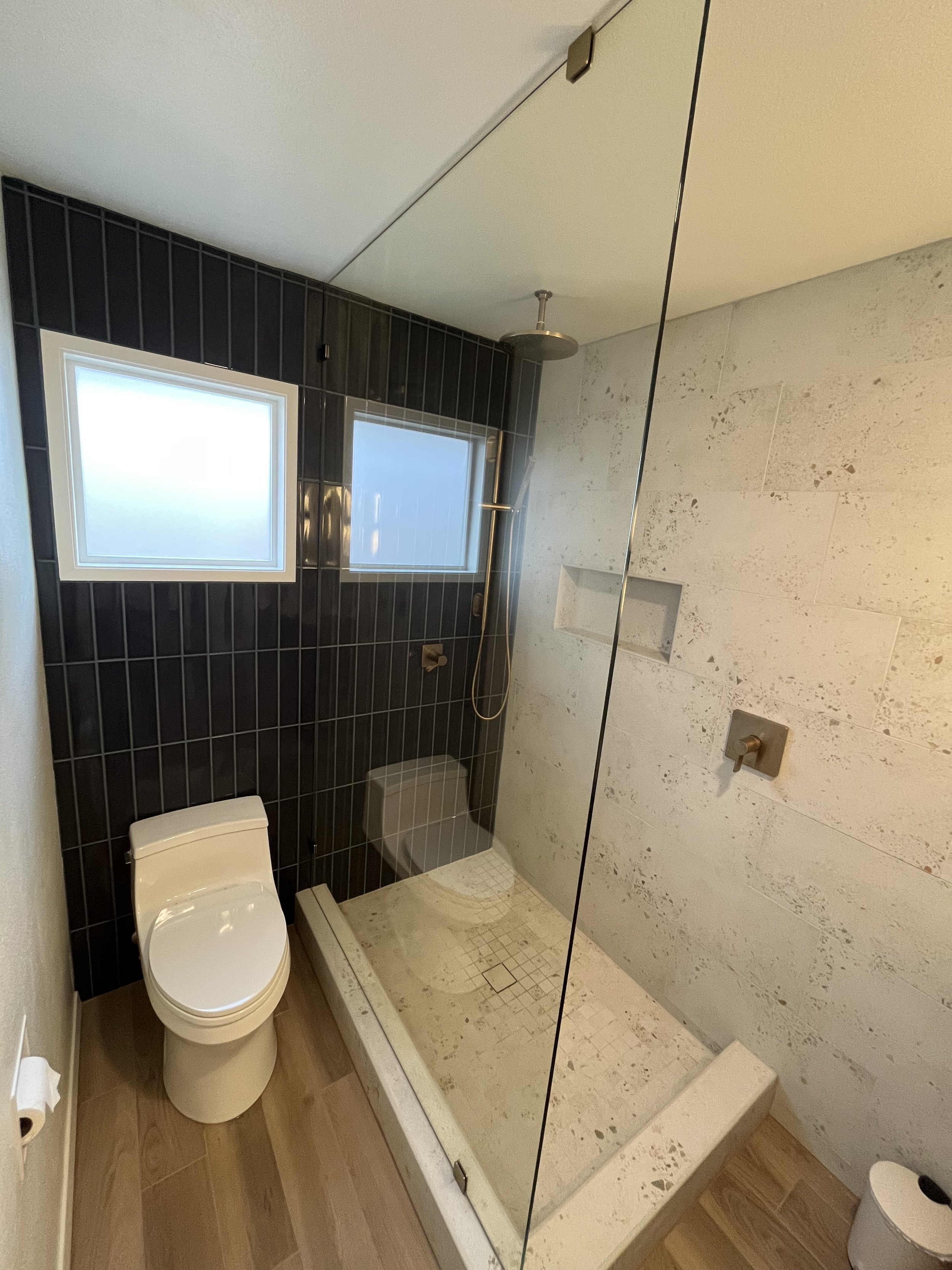 Modern bathroom with a walk-in shower featuring black tile wall and beige tile floor, white toilet, and frosted windows.