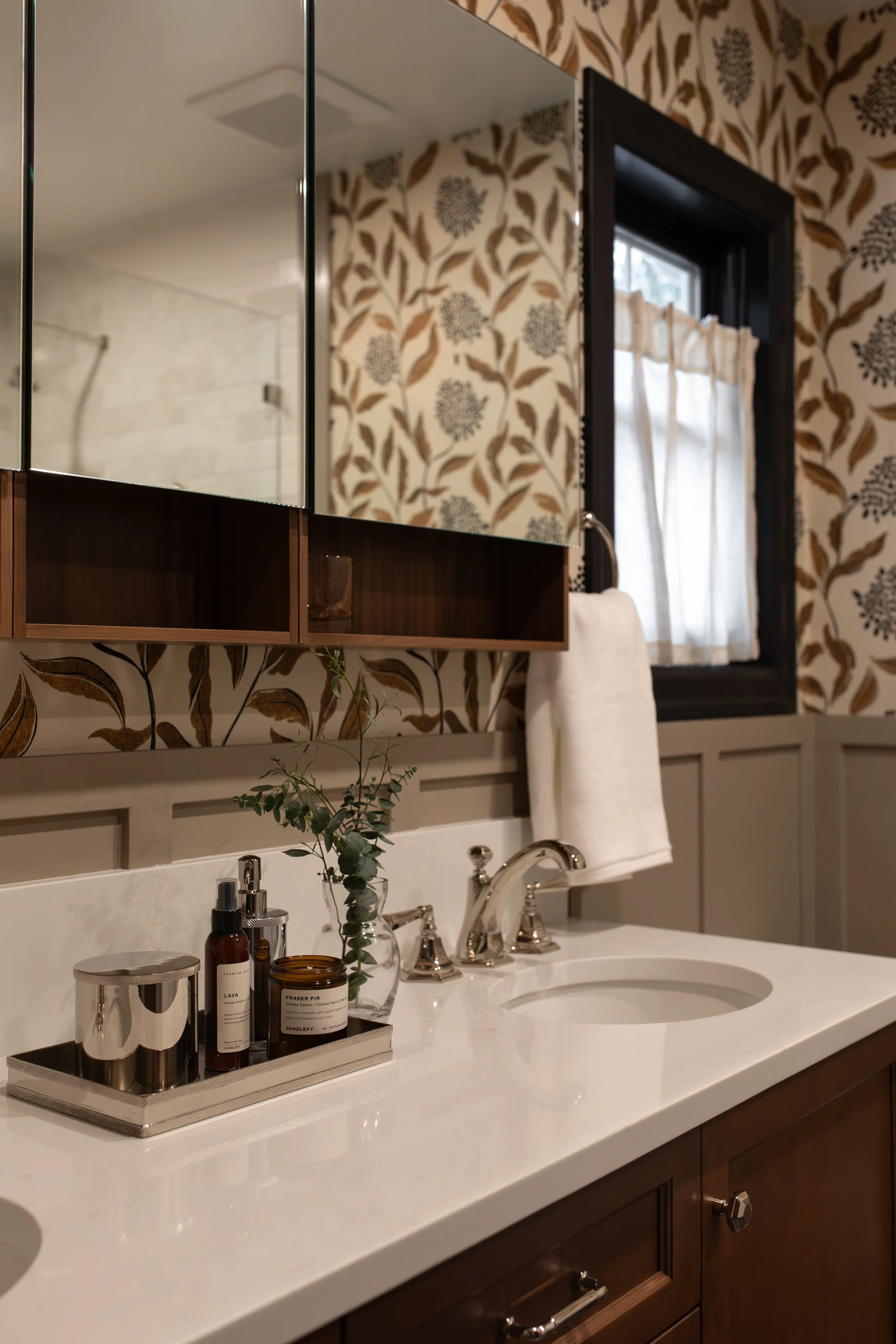 Bathroom vanity with mirror, soap dispenser, jars, small plant, towel, window, and floral wallpaper.