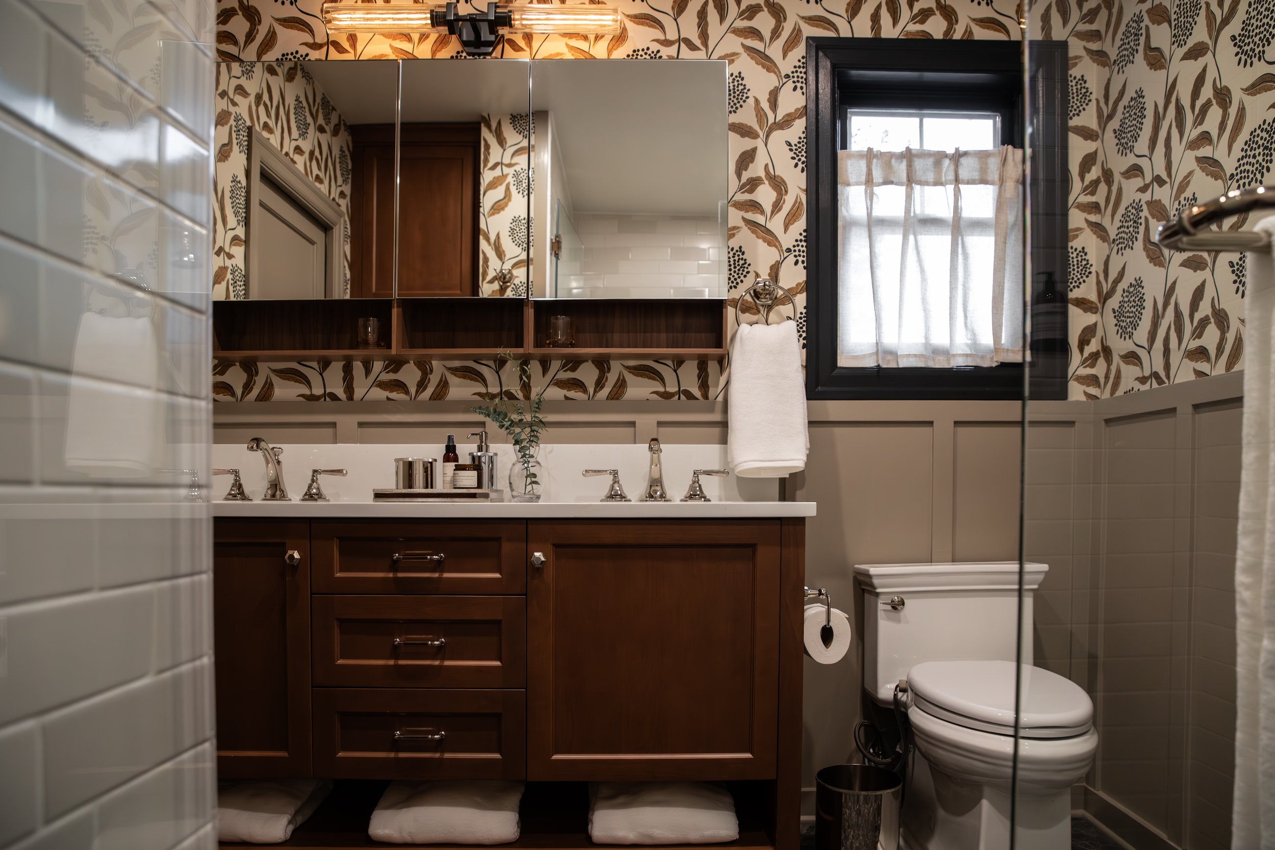 Bathroom with wooden vanity, white countertop, mirrors, framed window, tiled wall and a toilet.
