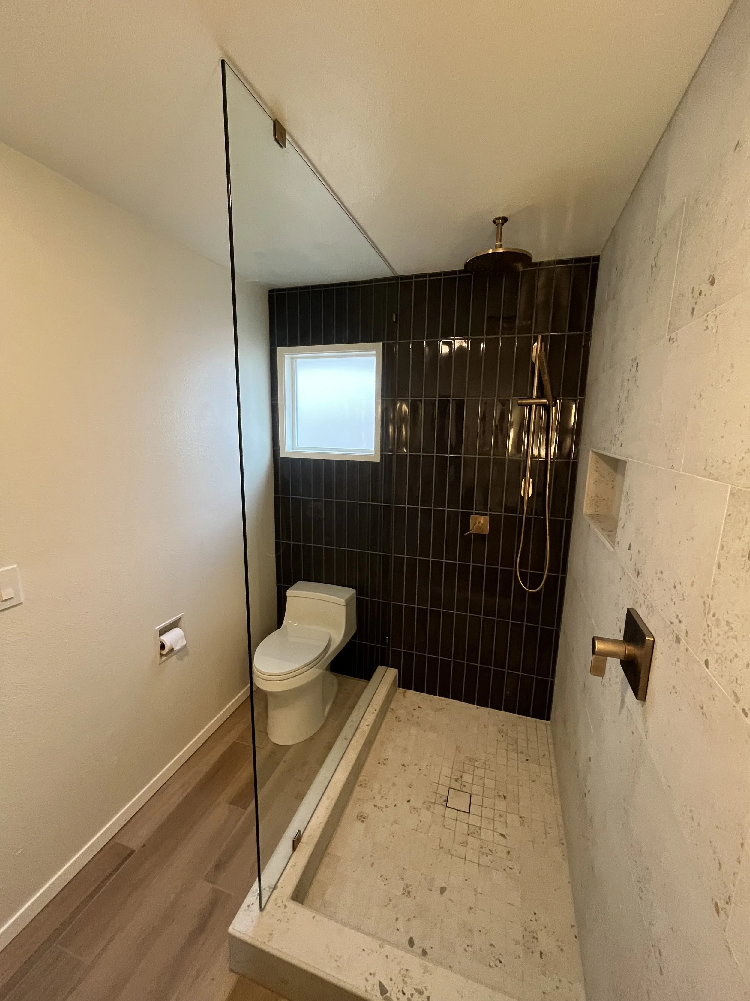 Bathroom with a toilet and a walk-in shower featuring black tiles on the wall, a small window, and a showerhead with a handheld attachment. The shower area has a low beige stone threshold and a built-in niche in the wall.