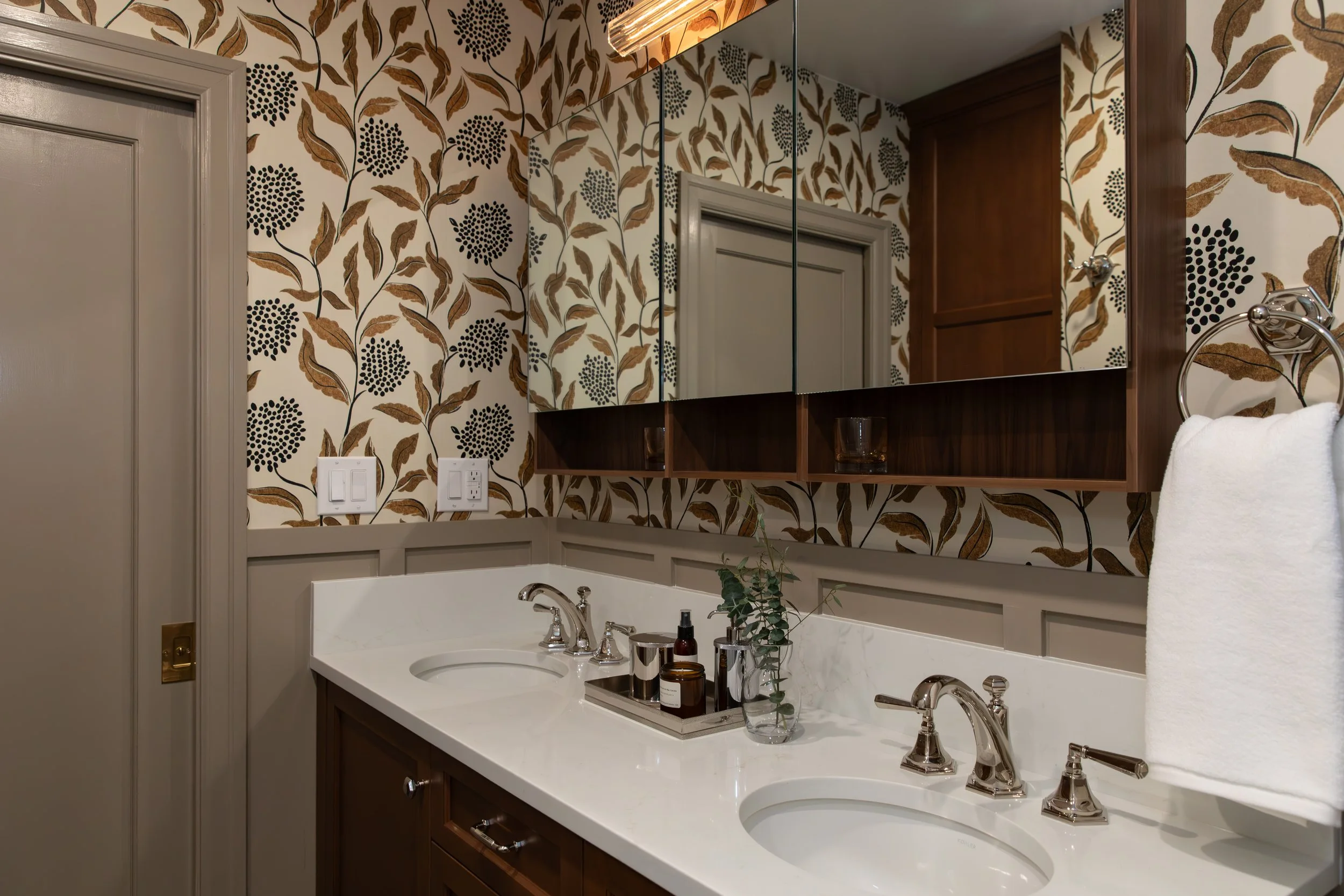 Bathroom vanity with double sinks, wood cabinetry, floral wallpaper, mirror cabinets, and a towel hanging on a ring