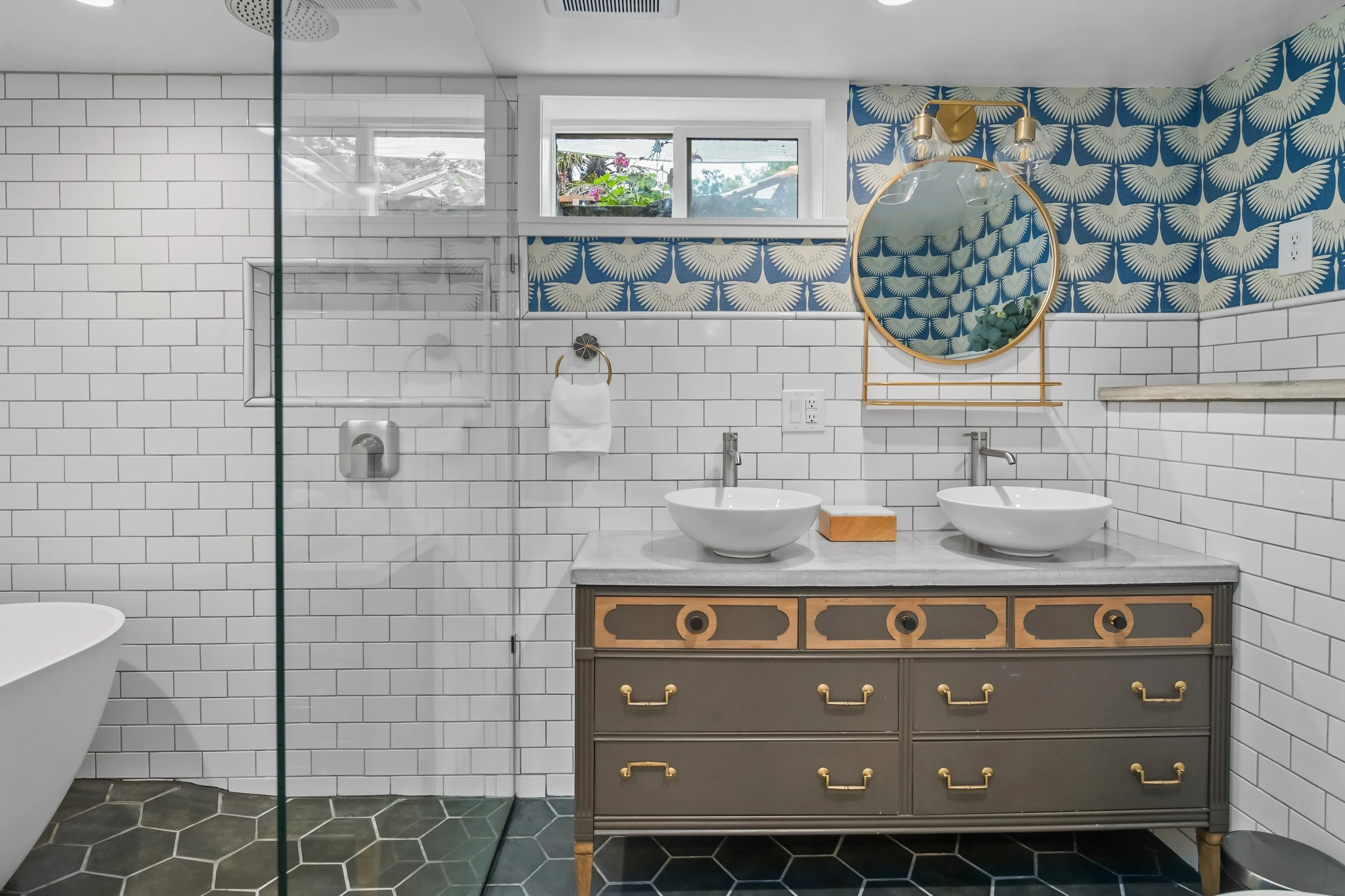 Bathroom with white subway tile walls, a double sink vanity with a gray and wood finish, a circular mirror with gold frame, blue wallpaper with a white leaf pattern, a small window, and a glass shower enclosure.
