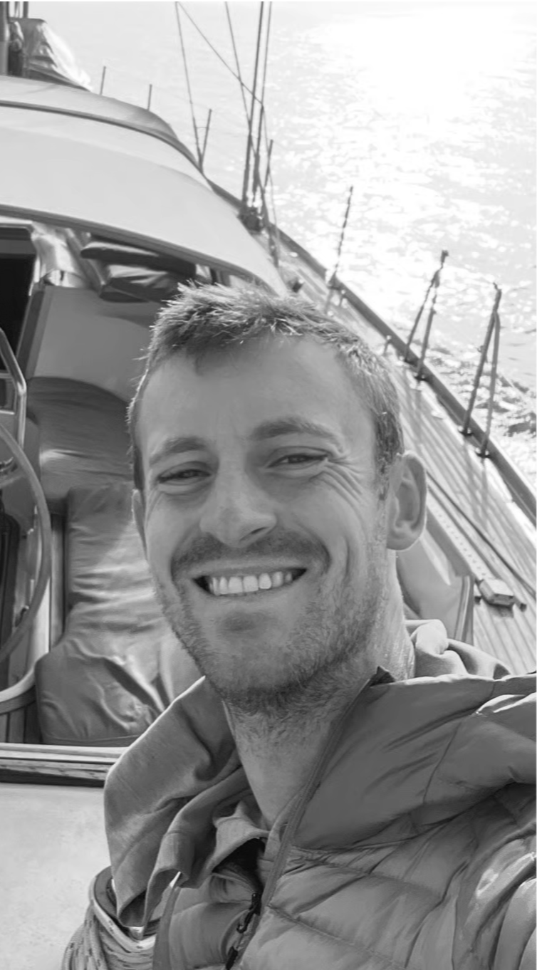A smiling man on a boat with water and sailboats in the background.