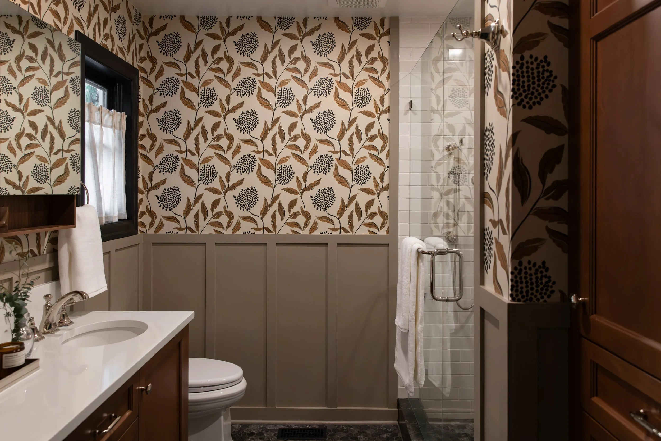 Bathroom with a white sink, toilet, shower with glass door, wooden cabinets, patterned wallpaper, and a window with a curtain.