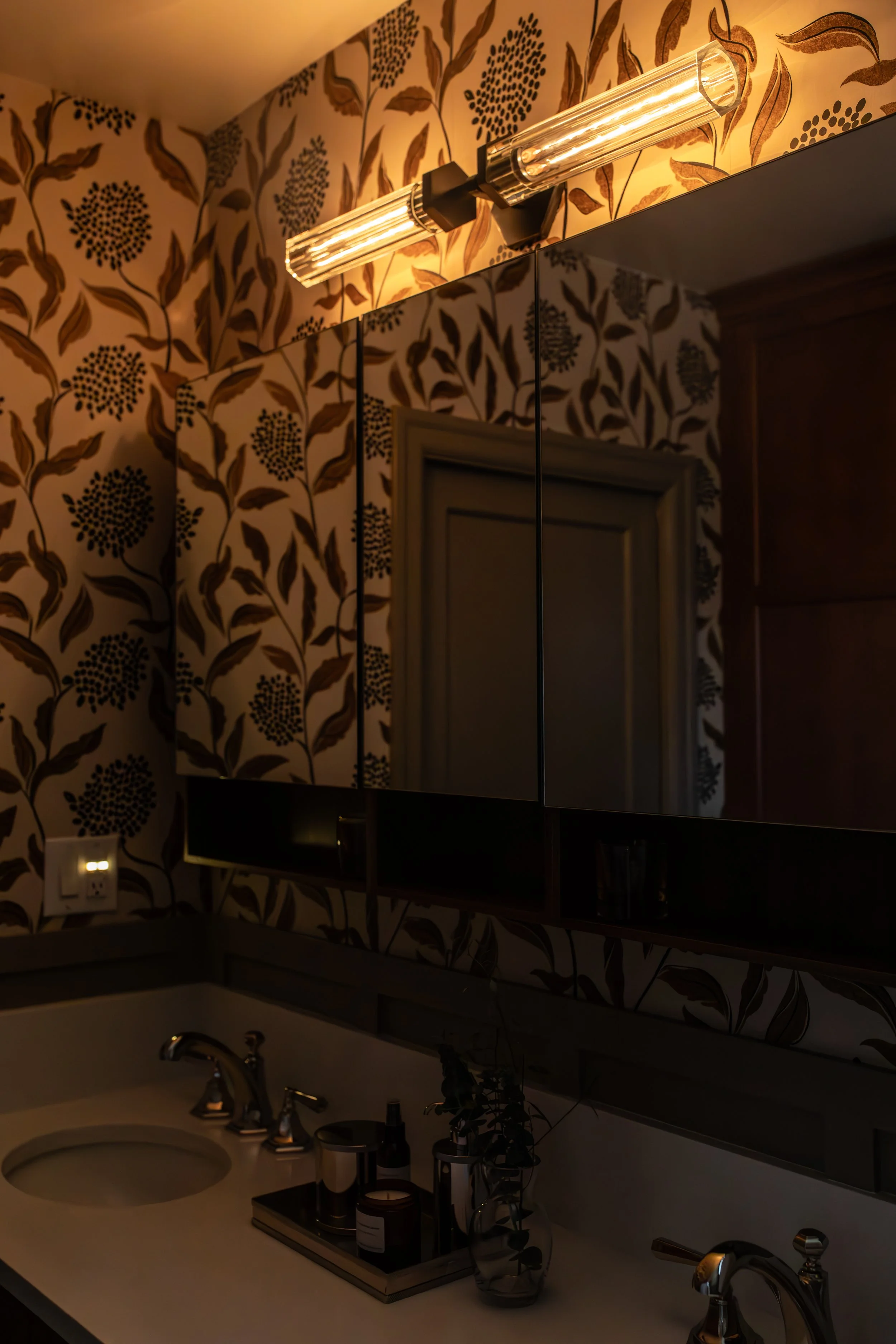 Bathroom vanity with a sink, mirror, wall-mounted light fixture, patterned wallpaper, vase with dried branches, and grooming products on a tray.