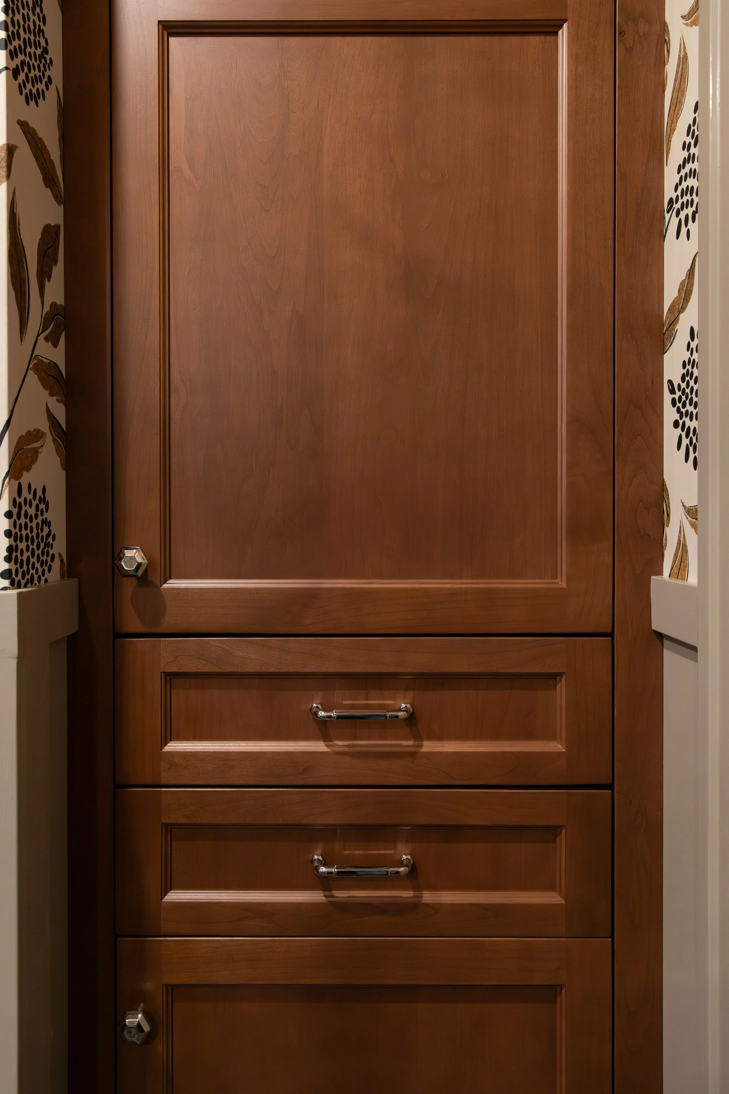 A tall wooden cabinet with three drawers and one door at the top, featuring metal handles, set against a wall with floral wallpaper.