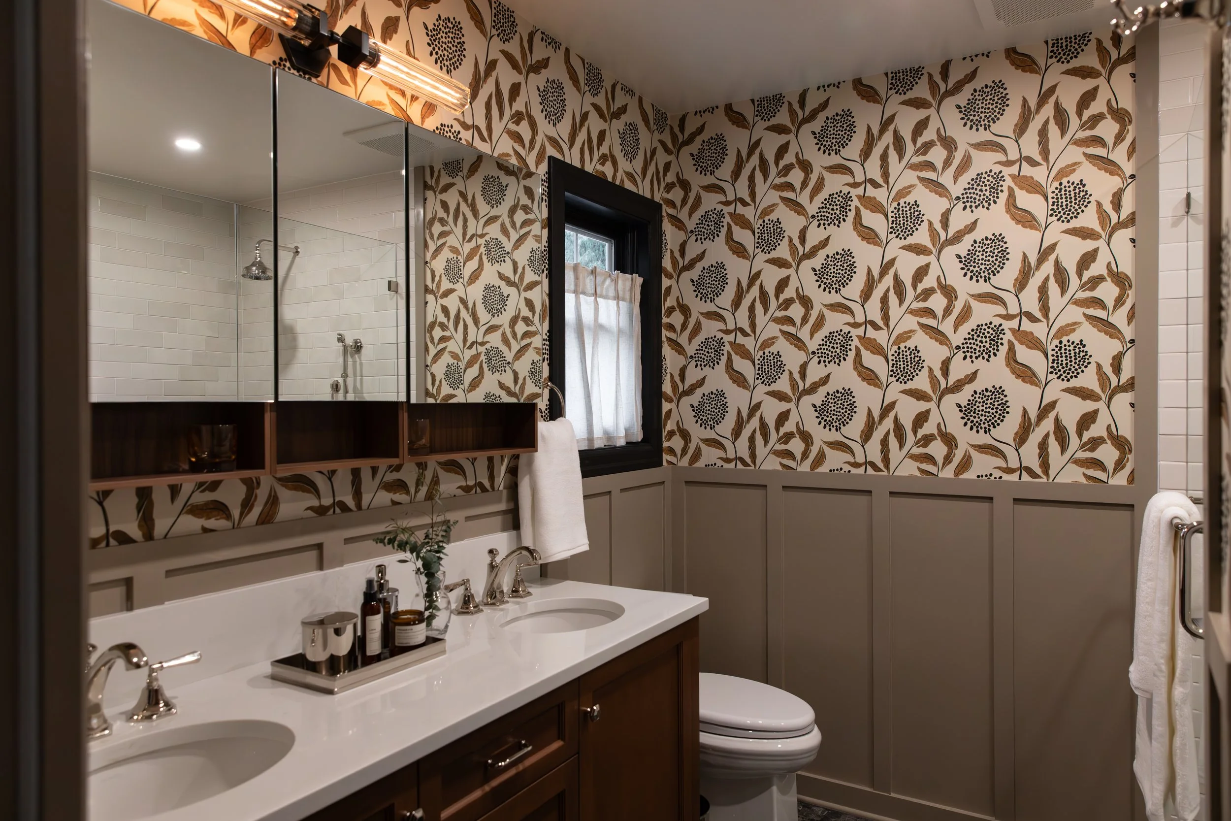 Bathroom with dual sinks, brown cabinetry, floral wallpaper, a window with curtains, and a mirror with lighting.