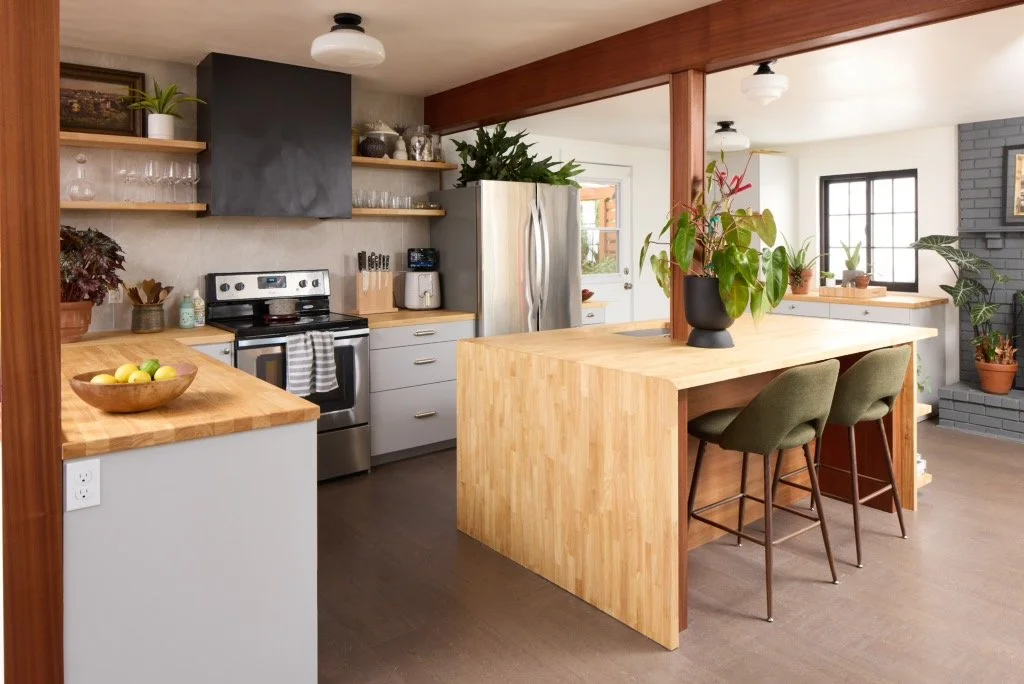 Modern kitchen with light wood accents, stainless steel appliances, open shelving with glassware and plants, including a large potted plant on the central island with two green bar stools, and large window with natural light.