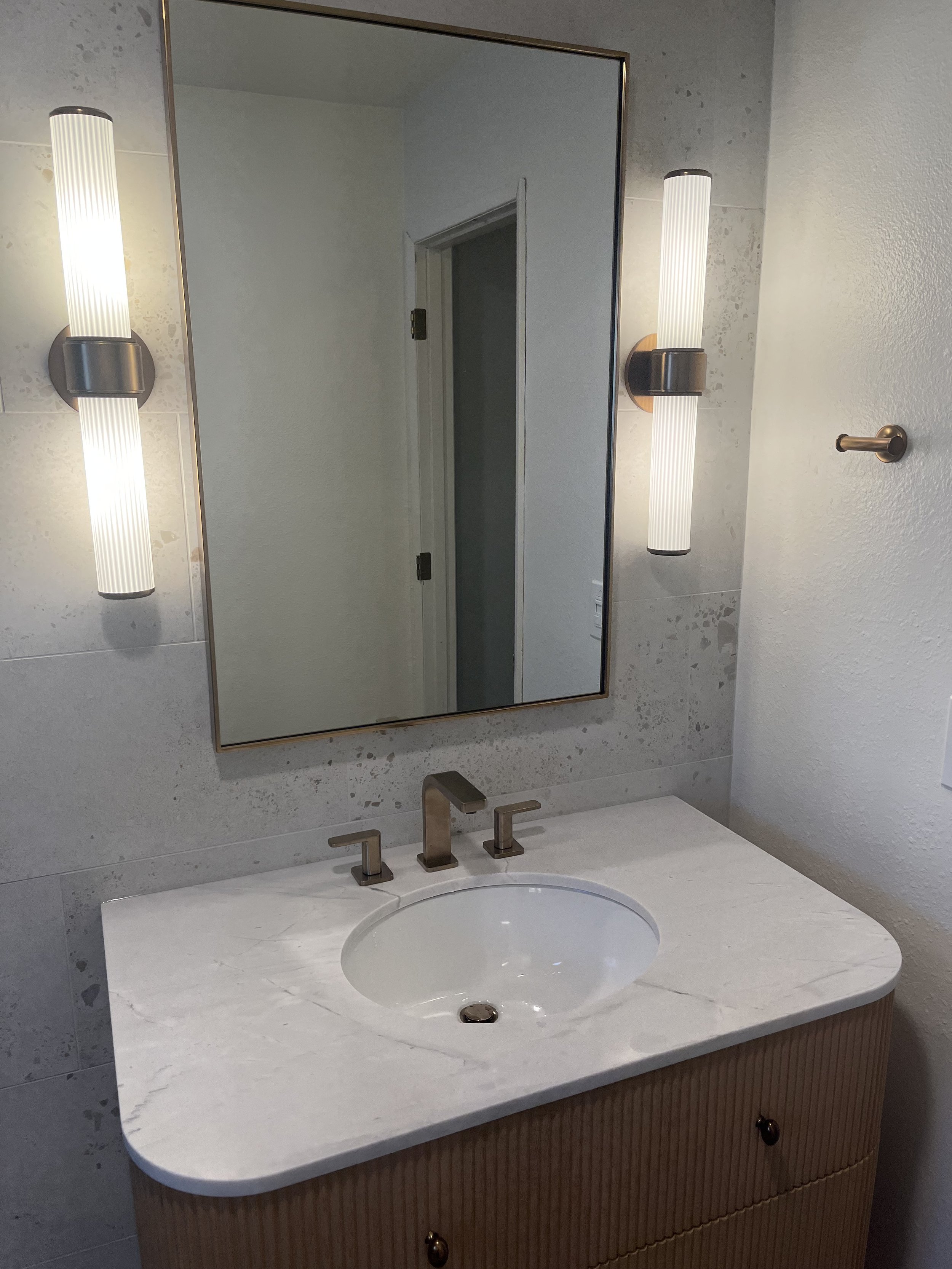 Bathroom vanity with a marble countertop, an oval sink, a modern faucet, and two wall-mounted lights on either side of a mirror.