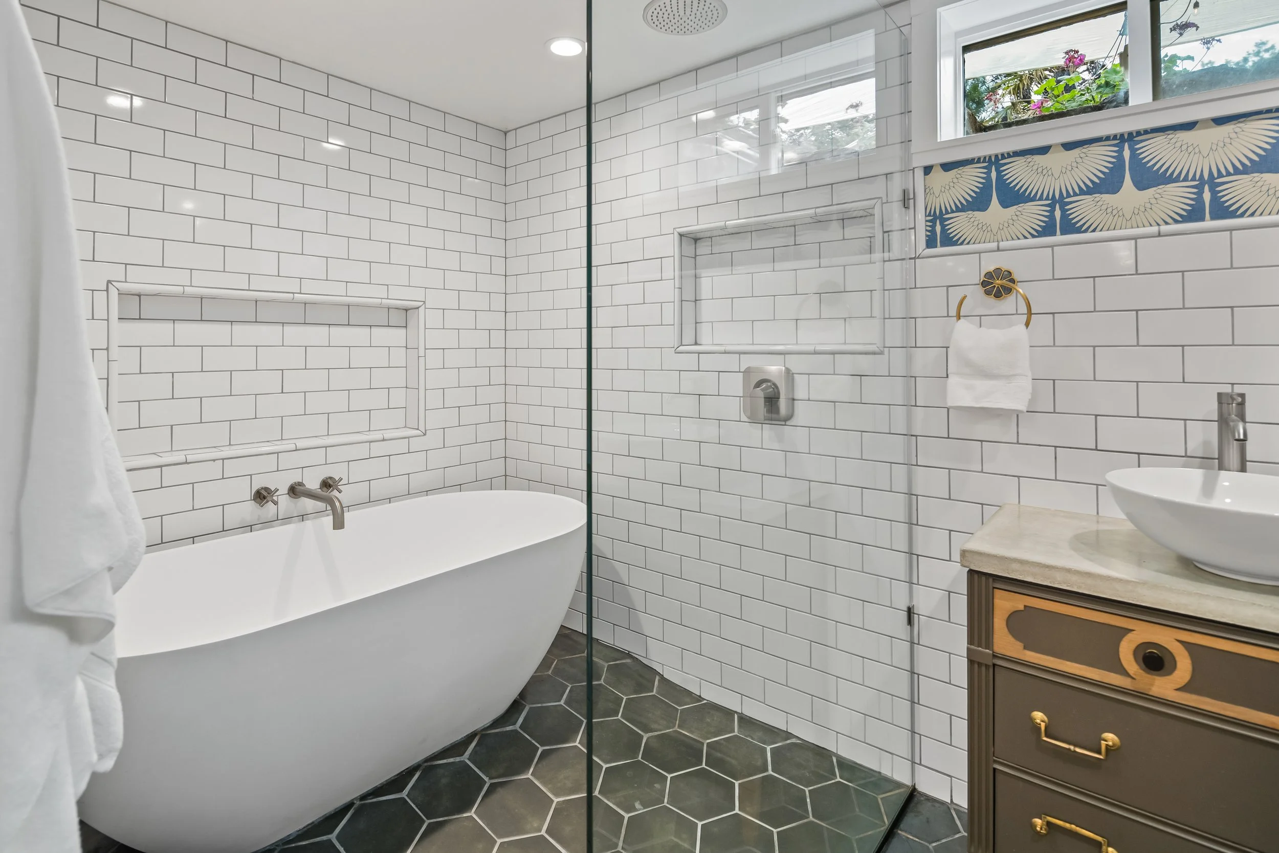Modern bathroom with white subway tile walls, a freestanding bathtub, a glass walk-in shower with built-in shelves, a small vanity with a vessel sink, and a window with floral and bird patterned valance.