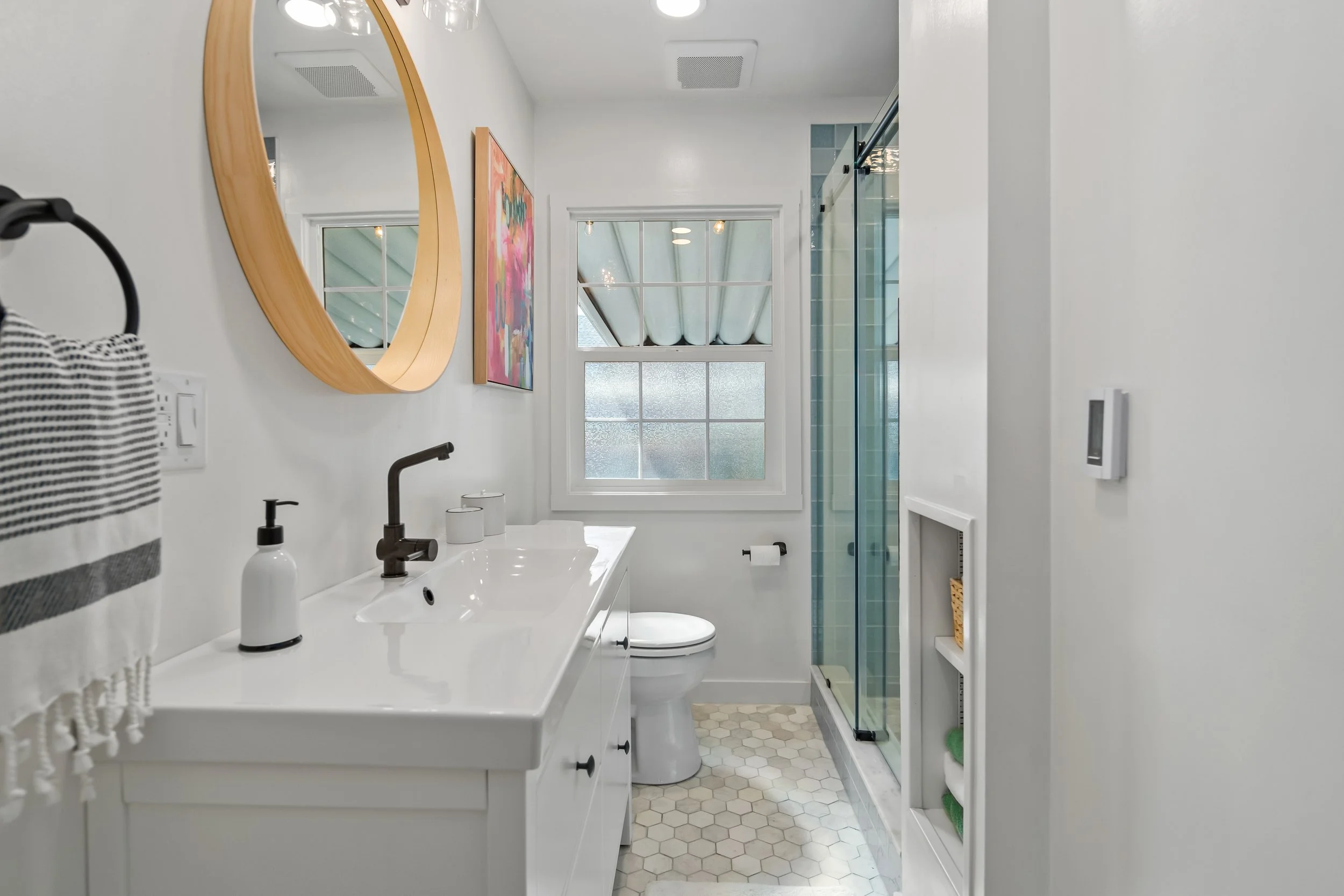 A modern bathroom with a white vanity, black faucet, oval mirror, colorful artwork, toilet, shower with glass doors, window, and built-in shelving with towels.