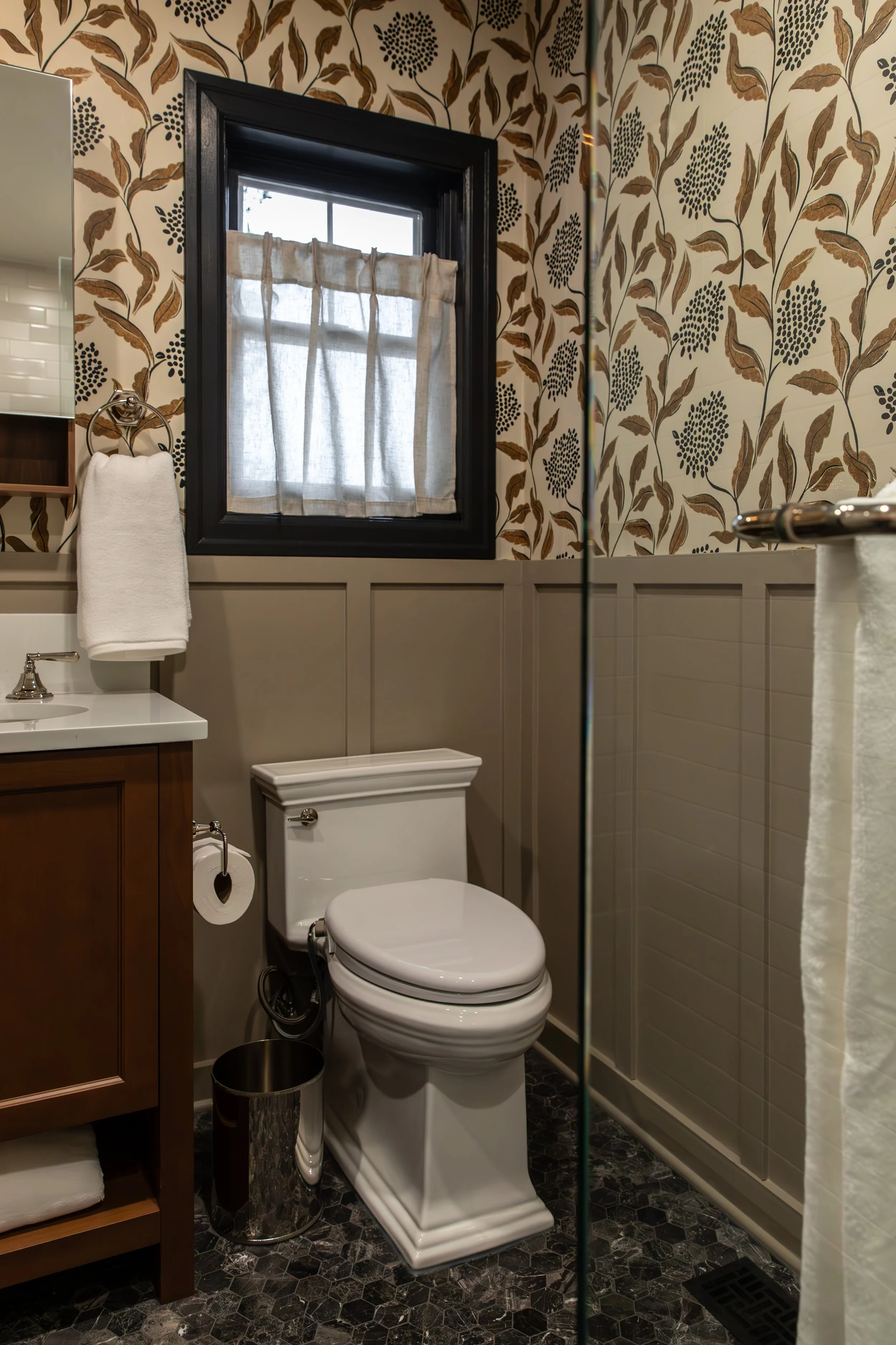 Bathroom with a white toilet, wooden vanity with a white sink, mirror, dark patterned tile floor, window with a small curtain, and floral wallpaper.