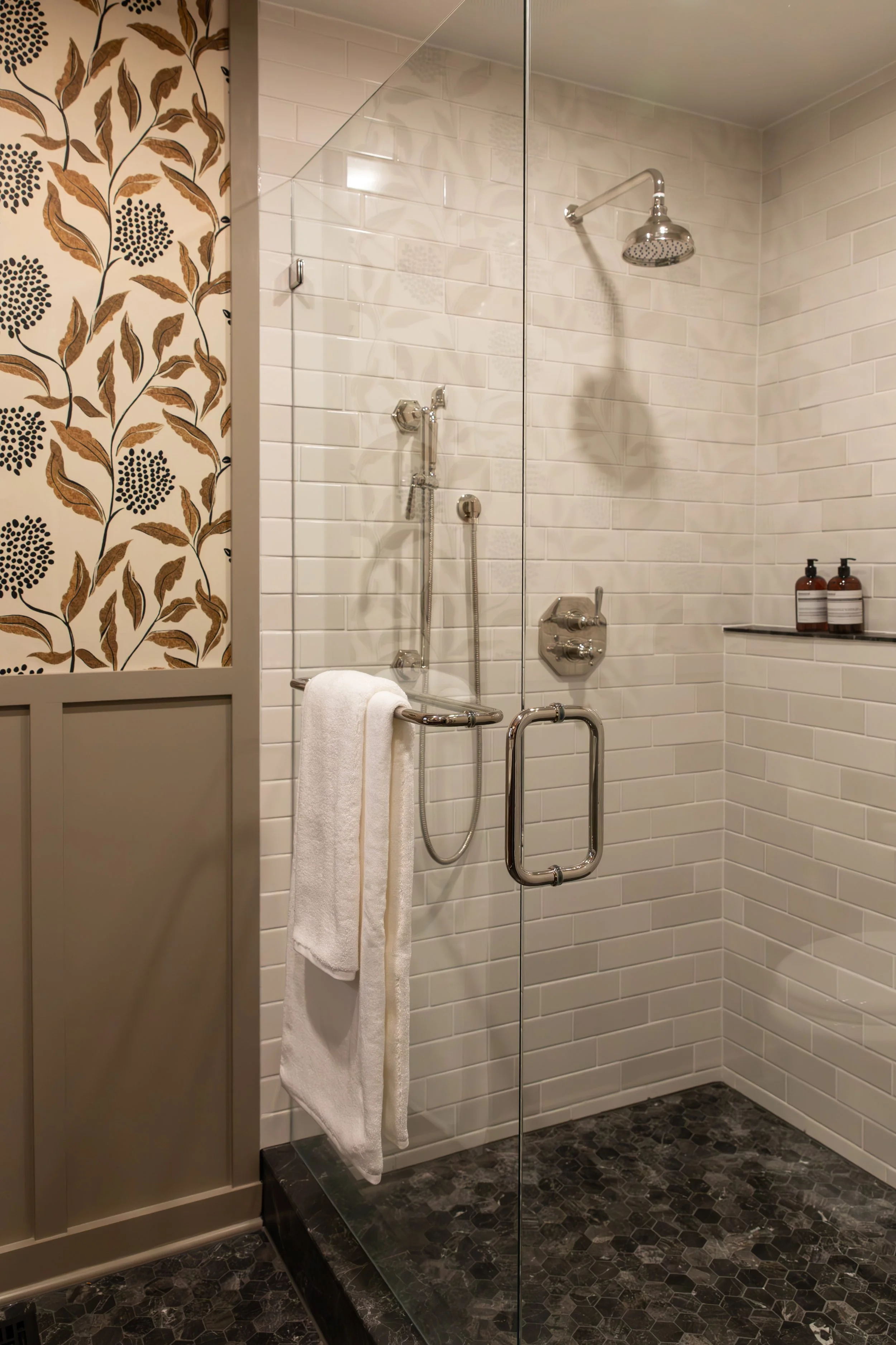 Modern shower with white subway tiles, a rain showerhead, a handheld shower, a glass door with a handle, a towel hanging on a bar, and two bottles of soap or shampoo on a shelf in a bathroom.