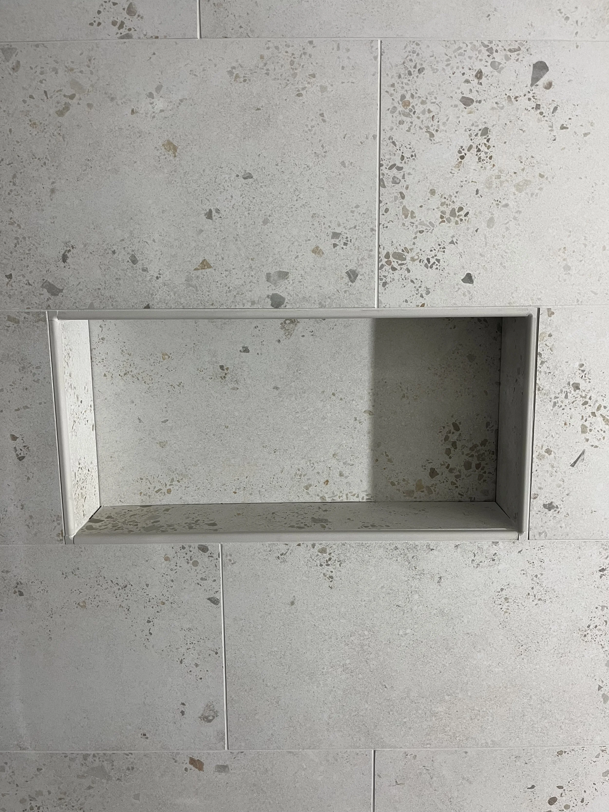 A rectangular wall niche with a concrete or stone back and sides, built into a gray, speckled tiled wall.
