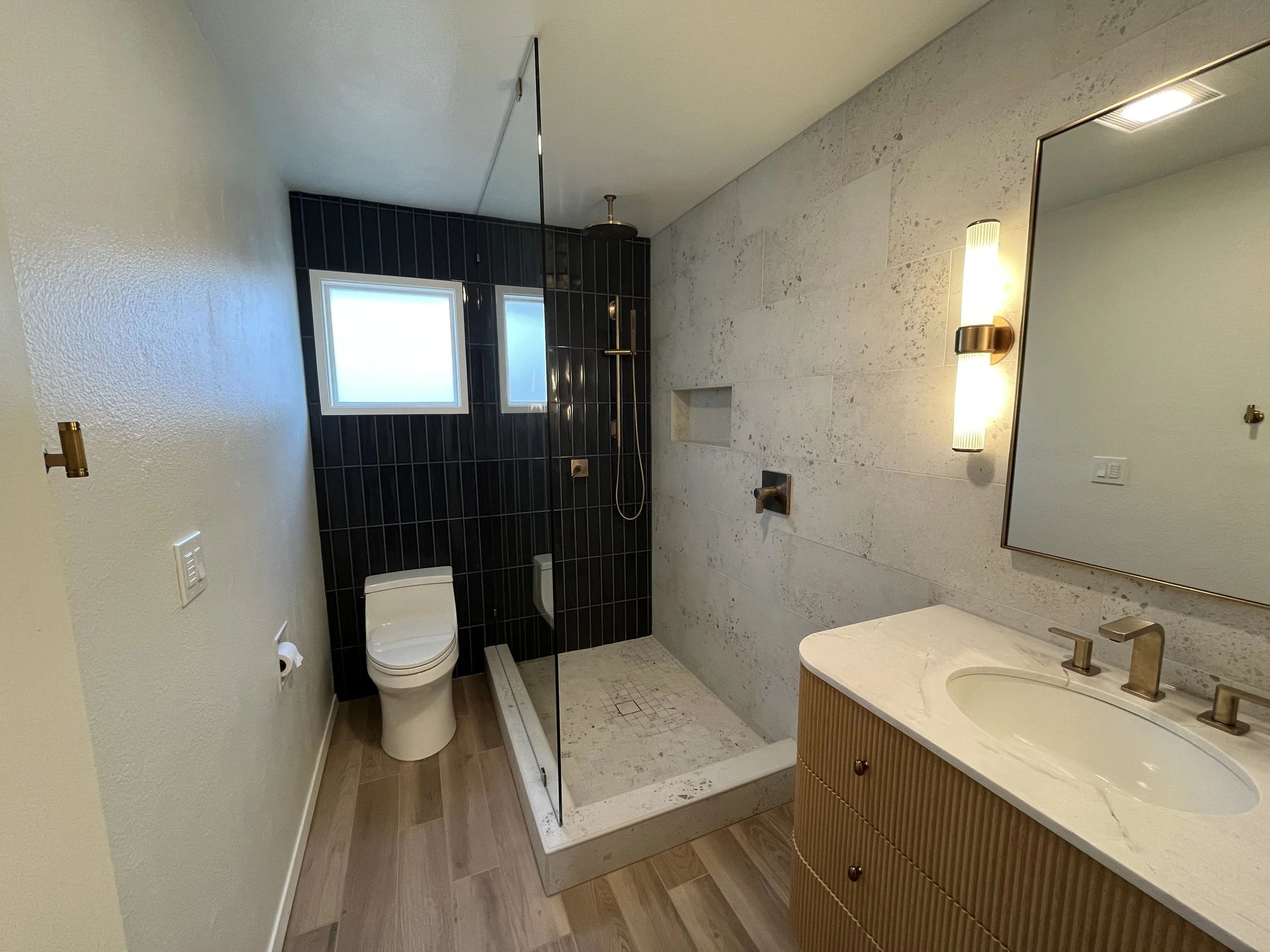 Modern bathroom featuring a walk-in shower with black tiles, a white toilet, a wooden vanity with a marble countertop, and a wall-mounted mirror with a vertical light fixture.